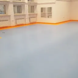 Jasnoniebieska, gładka podłoga sportowa z pomarańczowymi obrzeżami w sali gimnastycznej. Widoczna siatka zabezpieczająca okna i szara metalowa drzwi.