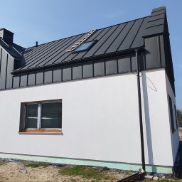 J&A POMAGAMY BUDOWAĆ - Nowoczesny dom jednorodzinny z białą elewacją i ciemnoszarą blachą na rąbek stojący, widoczny system rynnowy i okno dachowe z drabinką.