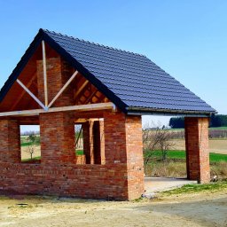 J&A POMAGAMY BUDOWAĆ - Surowa konstrukcja domu z czerwonej cegły z ciemnoszarą dachówką, widoczne drewniane elementy konstrukcyjne dachu, w tle pola i las, błękitne niebo.