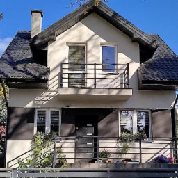 Dwupiętrowy dom z balkonem, ciemną dachówką i elementami elewacji w kolorze grafitowym, otoczony zielenią i ogrodzeniem.