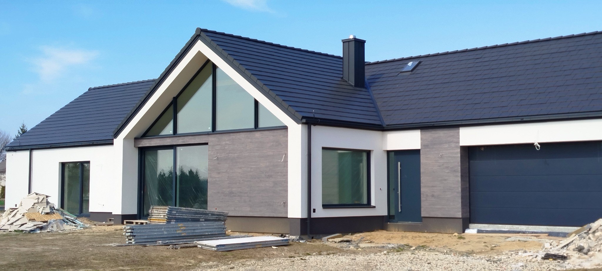 Nowoczesny dom jednorodzinny z czarną dachówką i dużymi oknami, w trakcie budowy, widoczne materiały budowlane na posesji.
