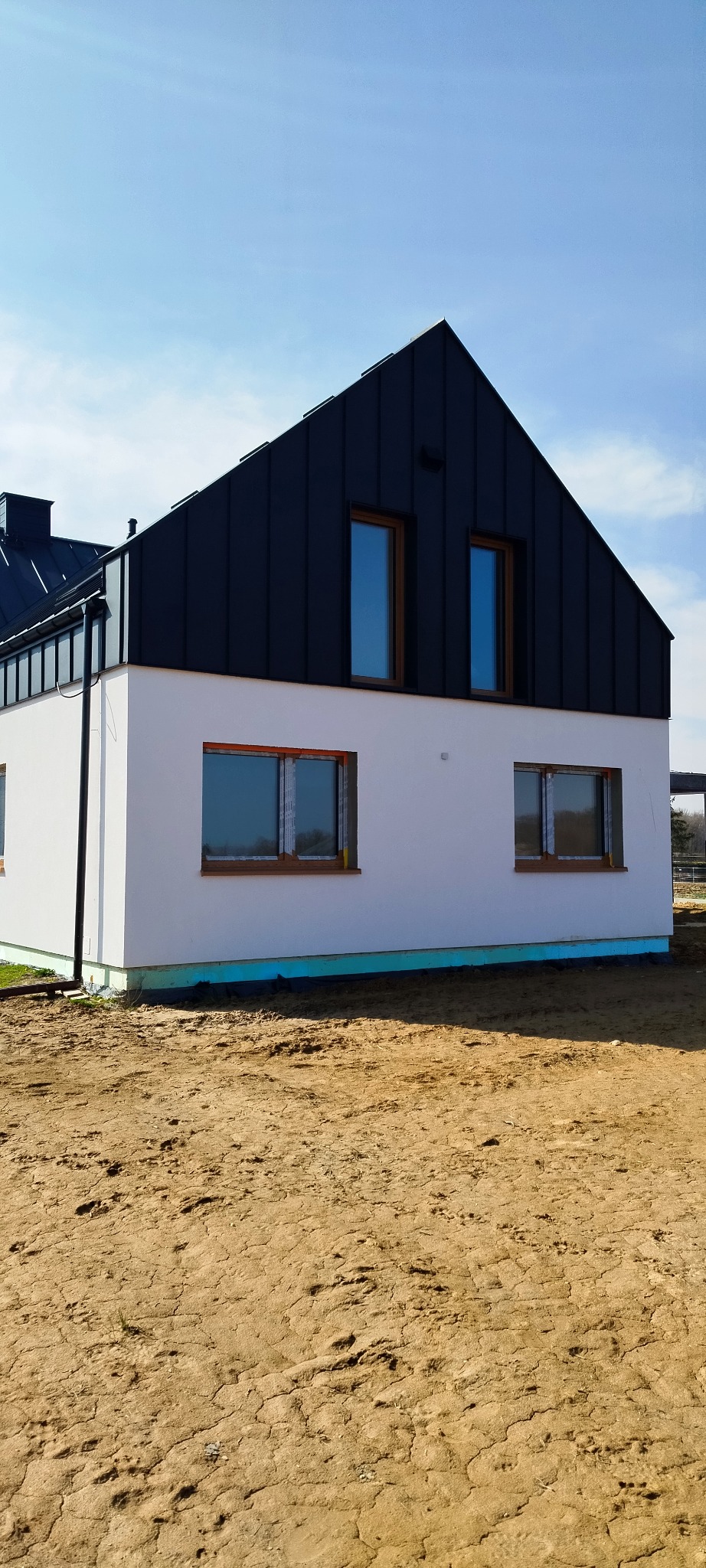 Nowoczesny dom jednorodzinny z czarnym dachem z blachy na rąbek stojący i białymi ścianami, widoczne okna z drewnianą stolarką, ocieplenie fundamentów i niezagospodarowany teren wokół.