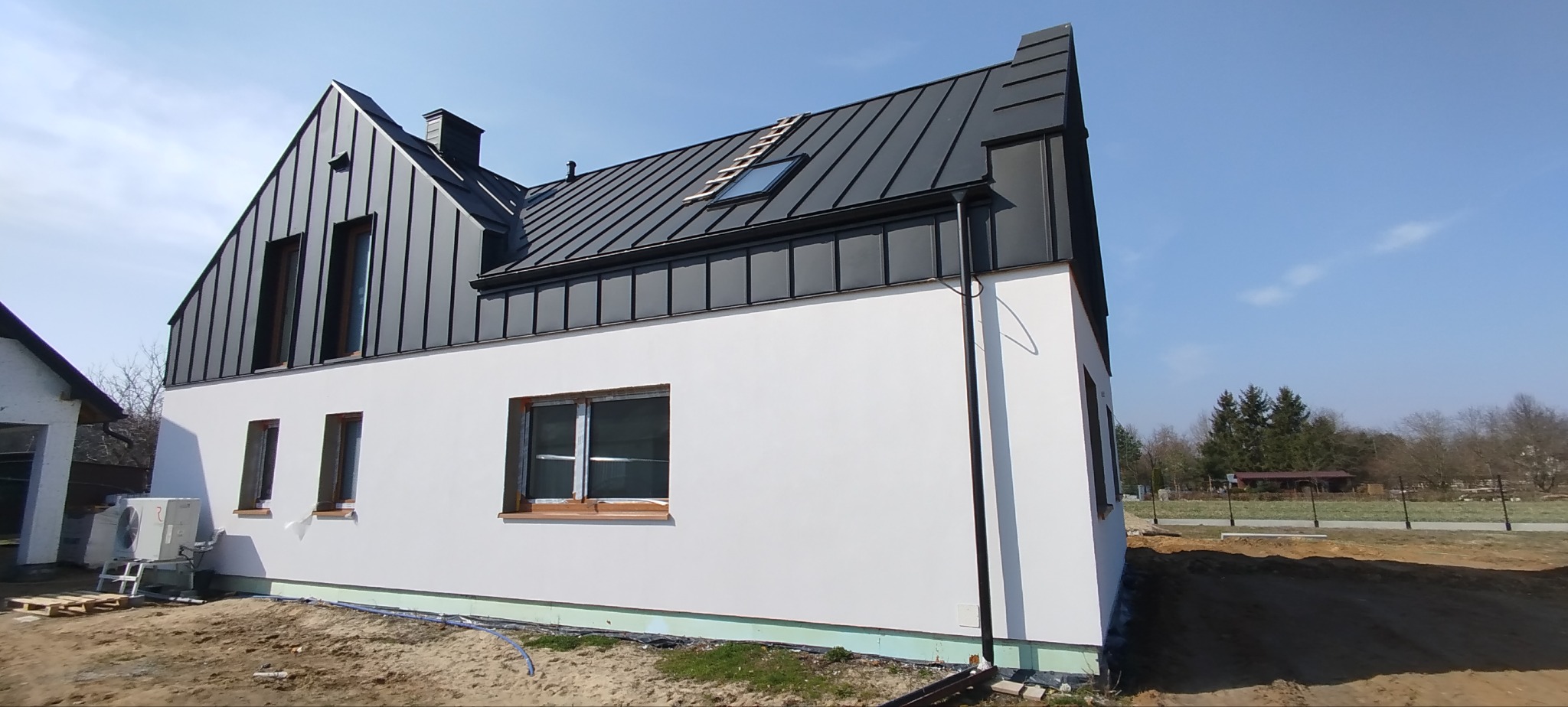 Nowoczesny dom jednorodzinny z białą elewacją i ciemnoszarą blachą na rąbek stojący, widoczny system rynnowy i okno dachowe z drabinką.