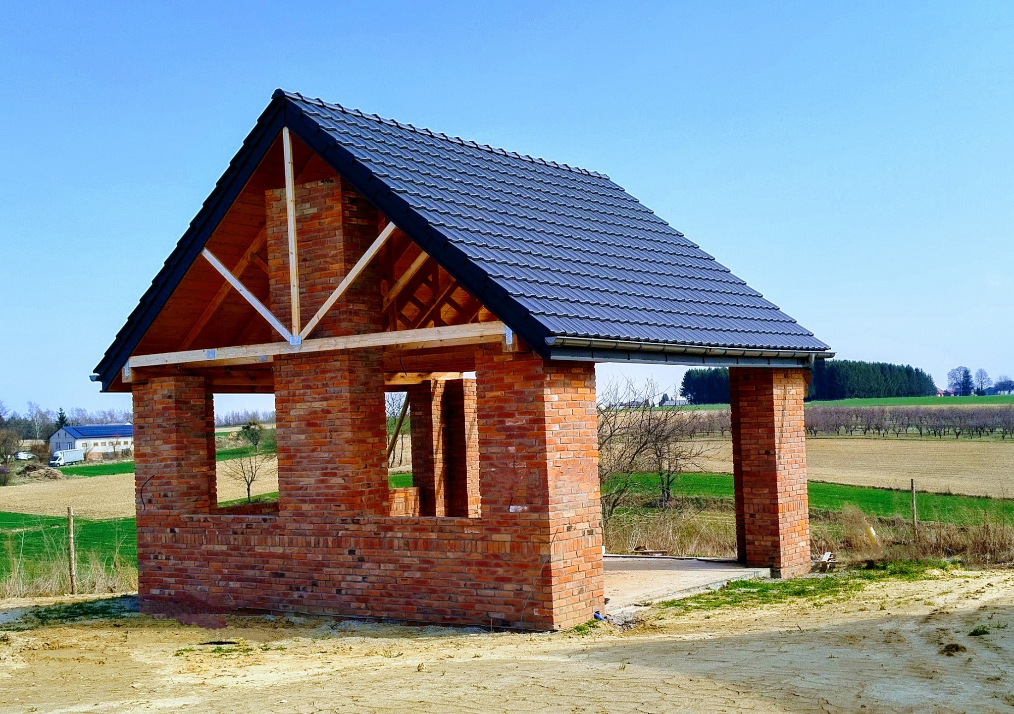 Surowa konstrukcja domu z czerwonej cegły z ciemnoszarą dachówką, widoczne drewniane elementy konstrukcyjne dachu, w tle pola i las, błękitne niebo.