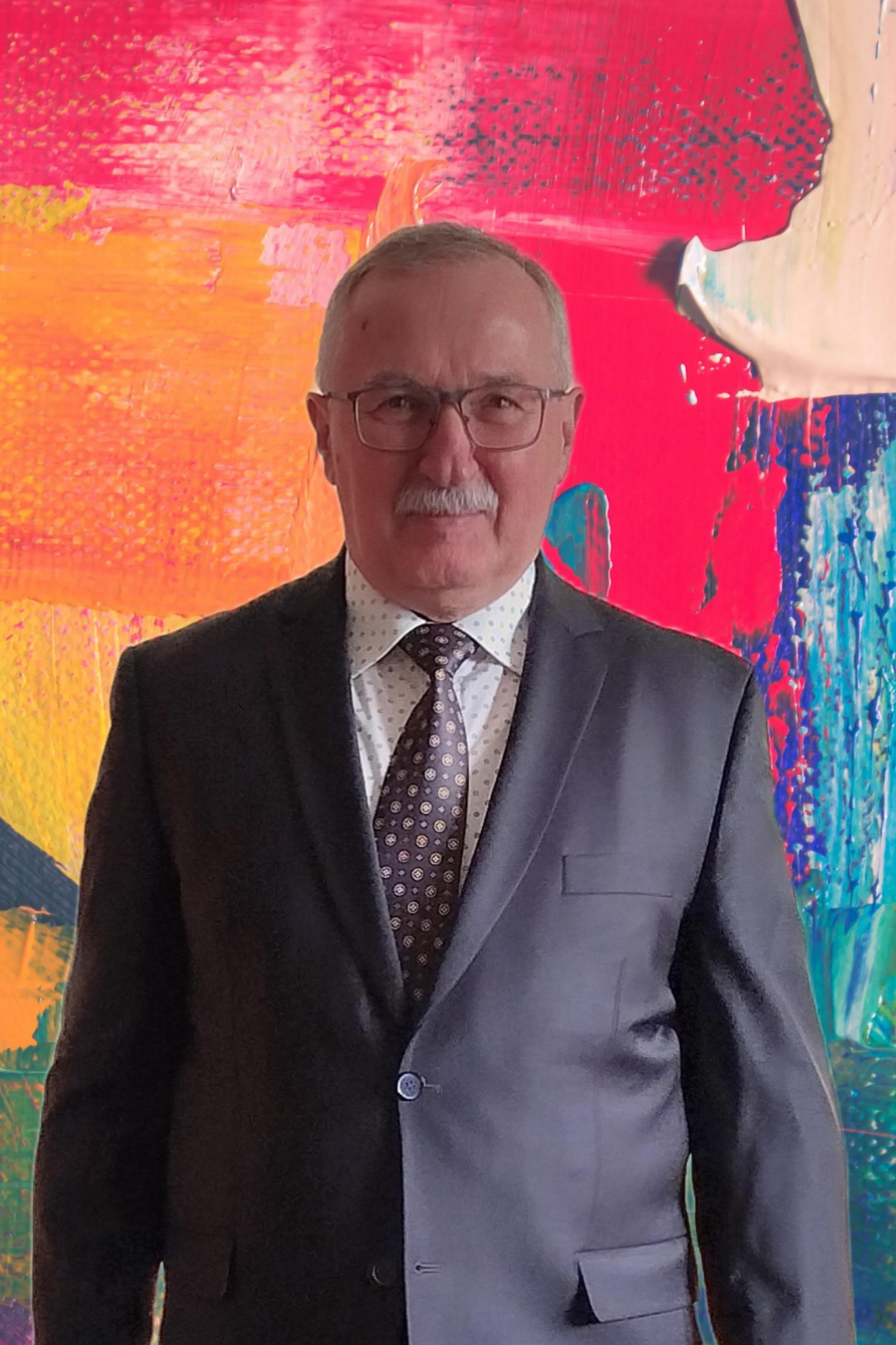 Portret mężczyzny w garniturze i okularach na tle abstrakcyjnego, kolorowego obrazu.