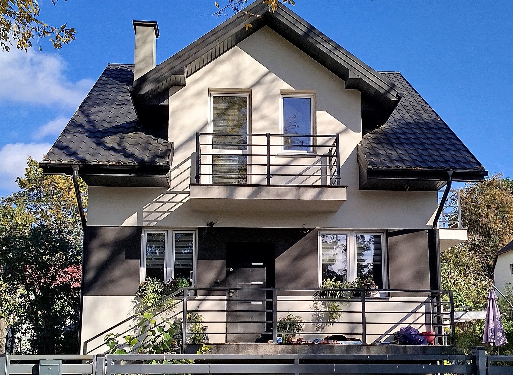 Dwupiętrowy dom z balkonem, ciemną dachówką i elementami elewacji w kolorze grafitowym, otoczony zielenią i ogrodzeniem.