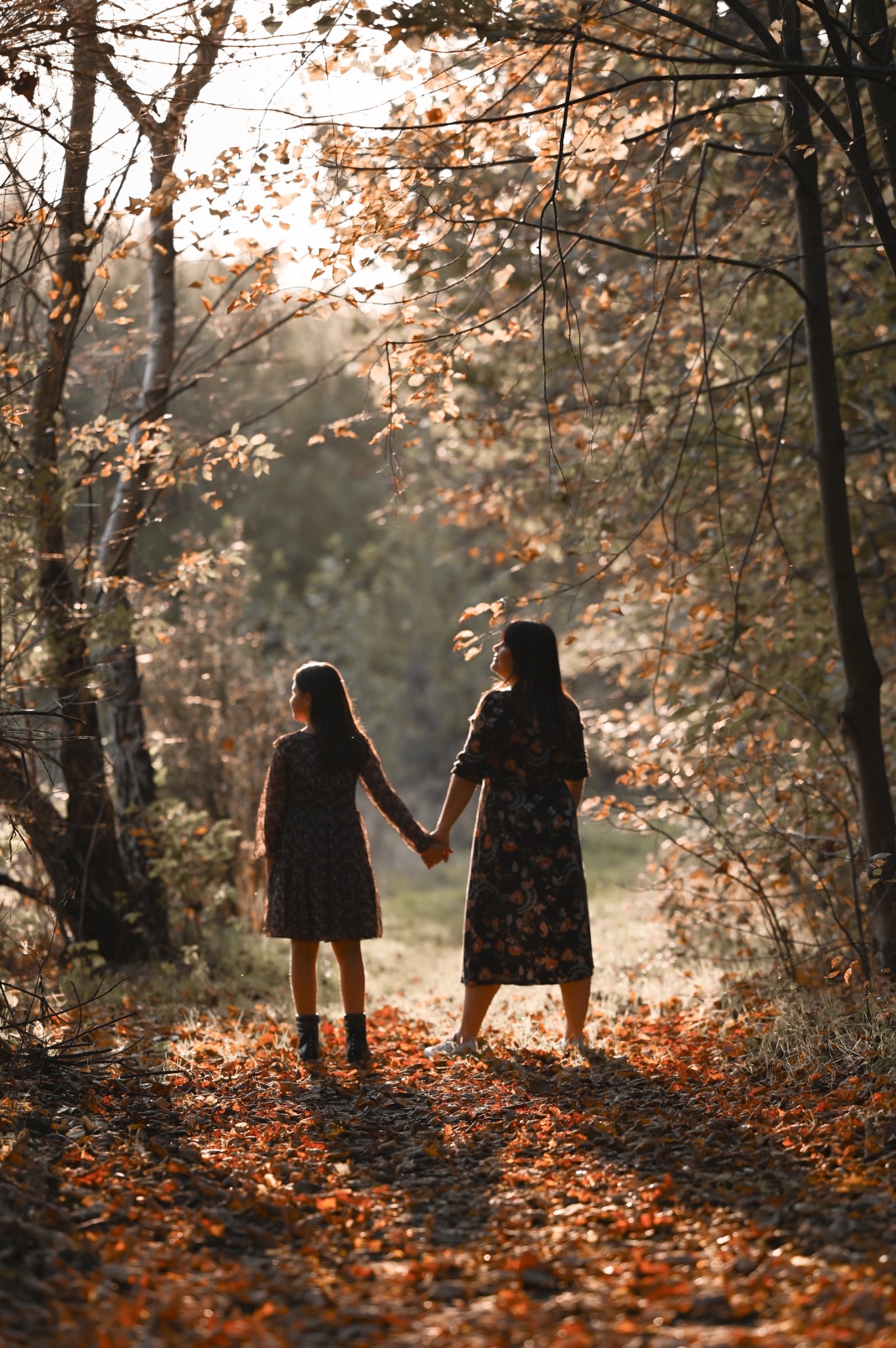Matka i córka trzymają się za ręce, spacerując po ścieżce w jesiennym lesie, oświetlonej promieniami słońca.
