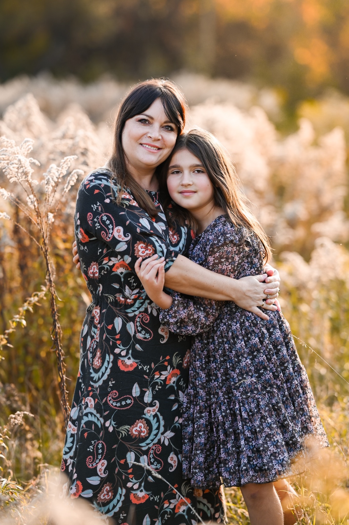 Matka i córka obejmujące się na tle łąki w ciepłym świetle zachodzącego słońca, obie ubrane w kwieciste sukienki.