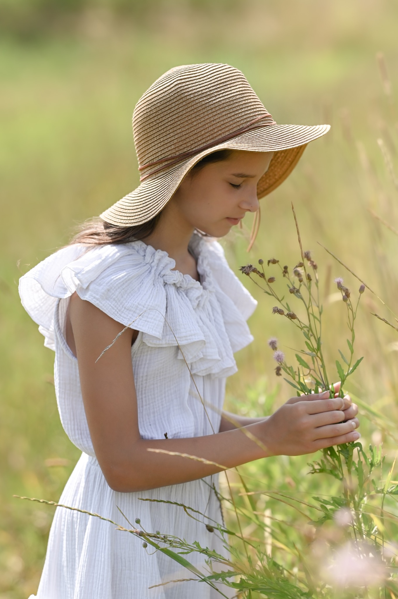 Dziewczynka w słomkowym kapeluszu i białej sukience, delikatnie dotykająca dłonią polnych kwiatów na łące w letnim słońcu.