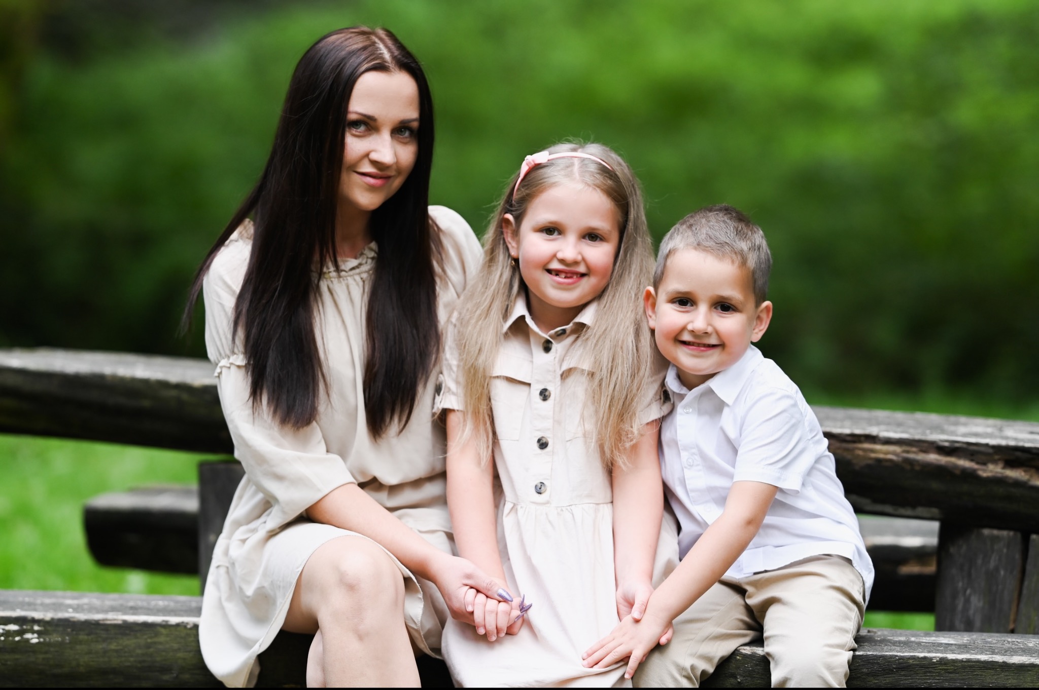 Kobieta z ciemnymi włosami siedzi na drewnianej ławce z dwójką uśmiechniętych dzieci, dziewczynką z różową opaską i chłopcem w białej koszuli, na tle zieleni.