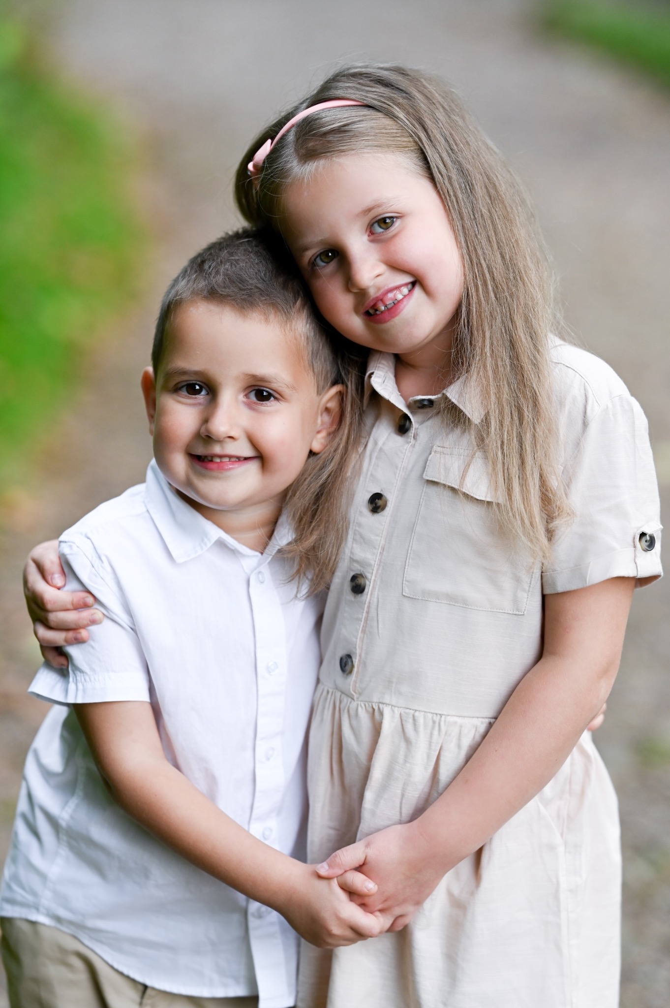 Uśmiechnięte rodzeństwo: chłopiec w białej koszuli i dziewczynka w beżowej sukience, obejmują się na zewnątrz, dziewczynka ma różową opaskę we włosach.