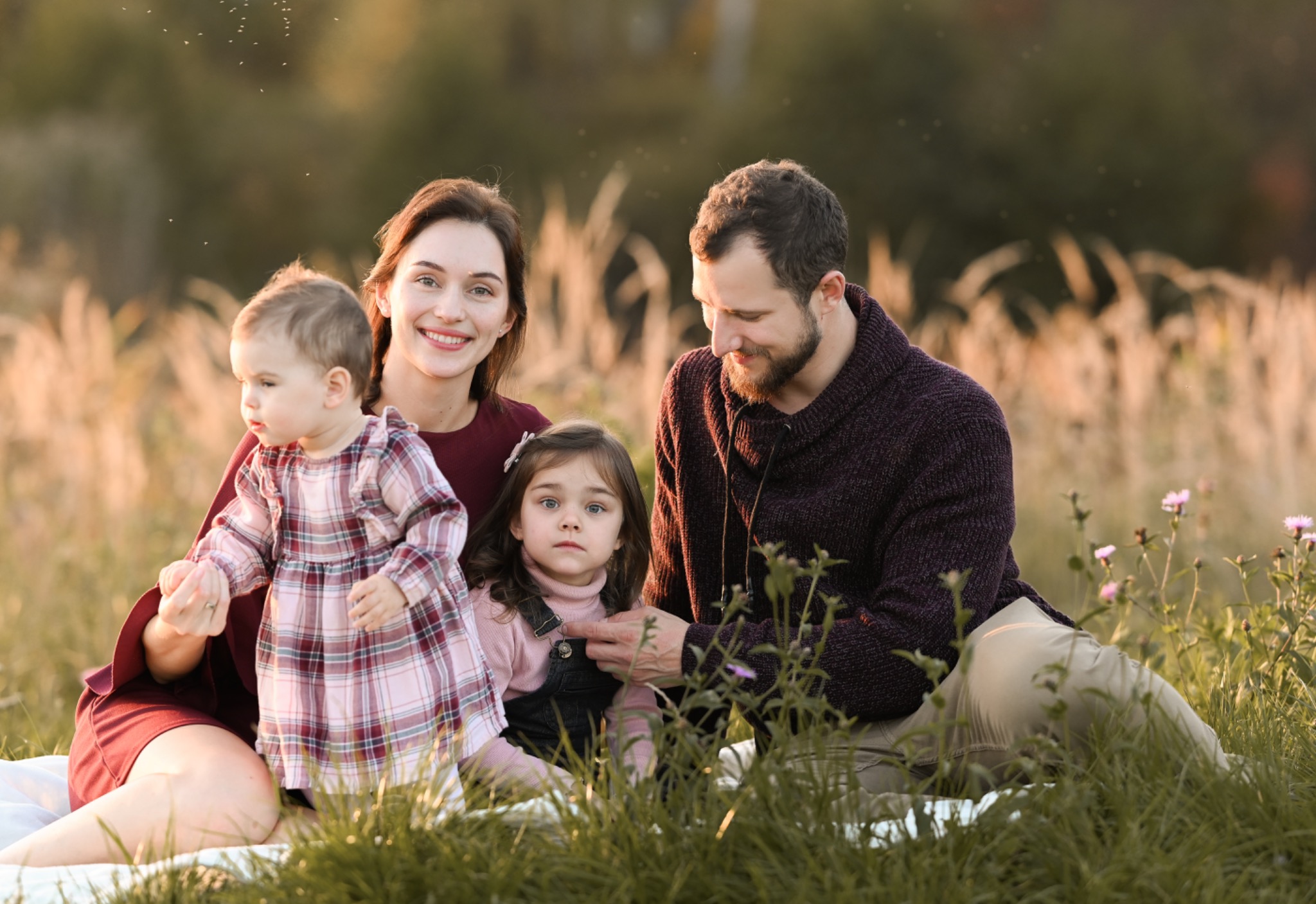 Rodzinne zdjęcie: matka z uśmiechem, ojciec z brodą i dwie córki na łące w promieniach zachodzącego słońca.