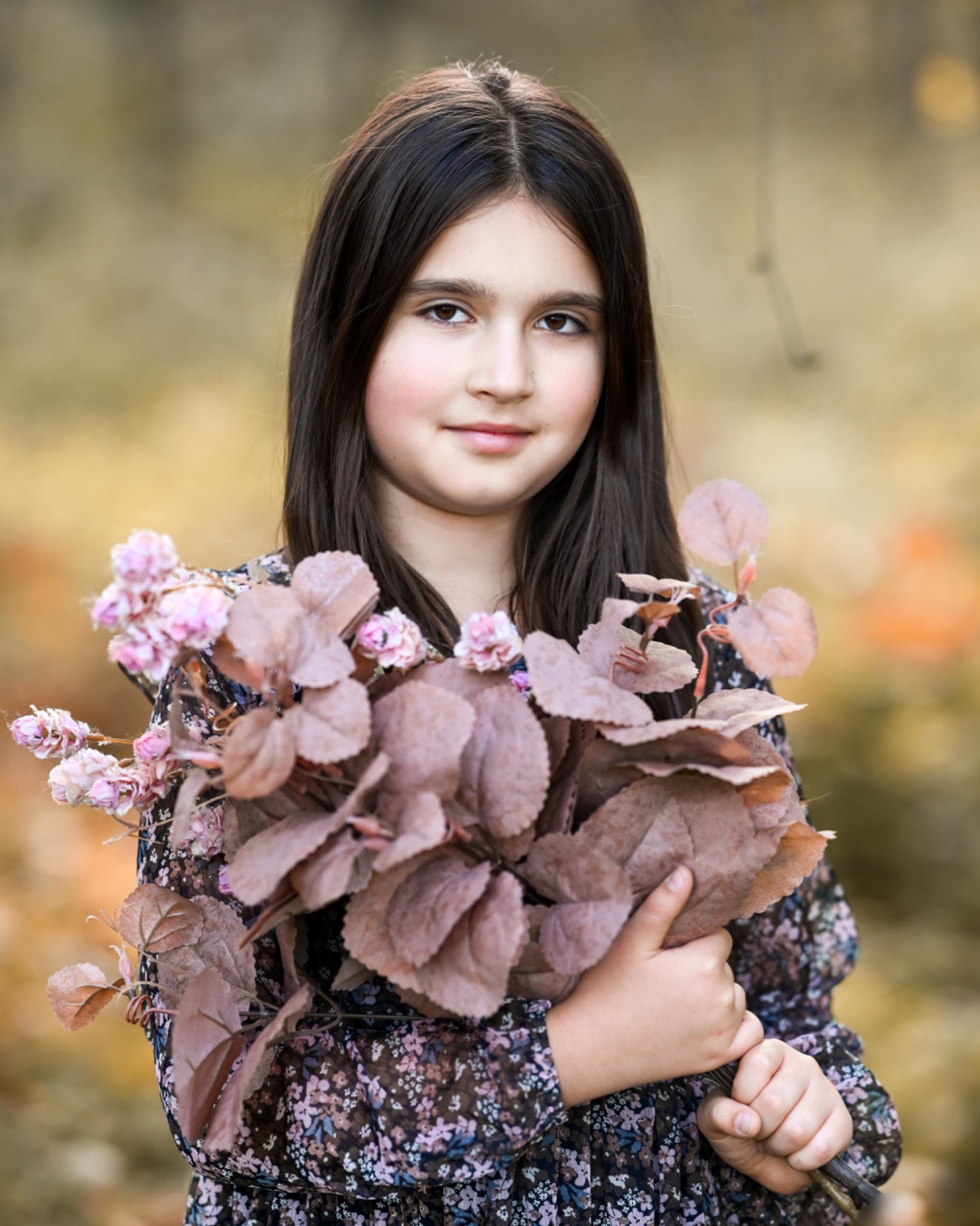 Portret uśmiechniętej dziewczynki z brązowymi włosami trzymającej bukiet brązowych liści i różowych kwiatów na tle rozmytej jesiennej scenerii.