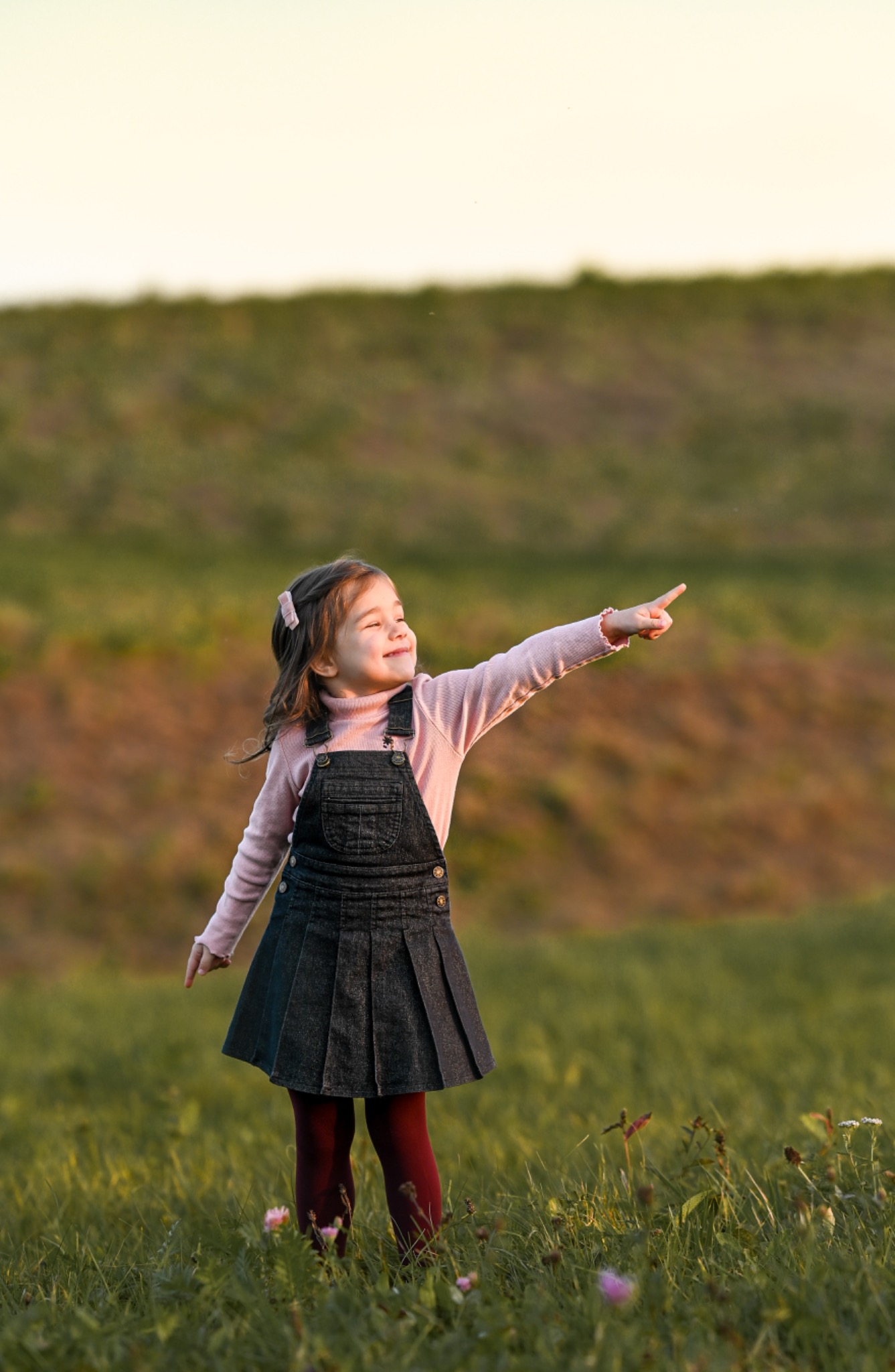 Dziewczynka w dżinsowej sukience i różowym swetrze stoi na łące, wyciągając rękę i wskazując coś palcem, z uśmiechem na twarzy.