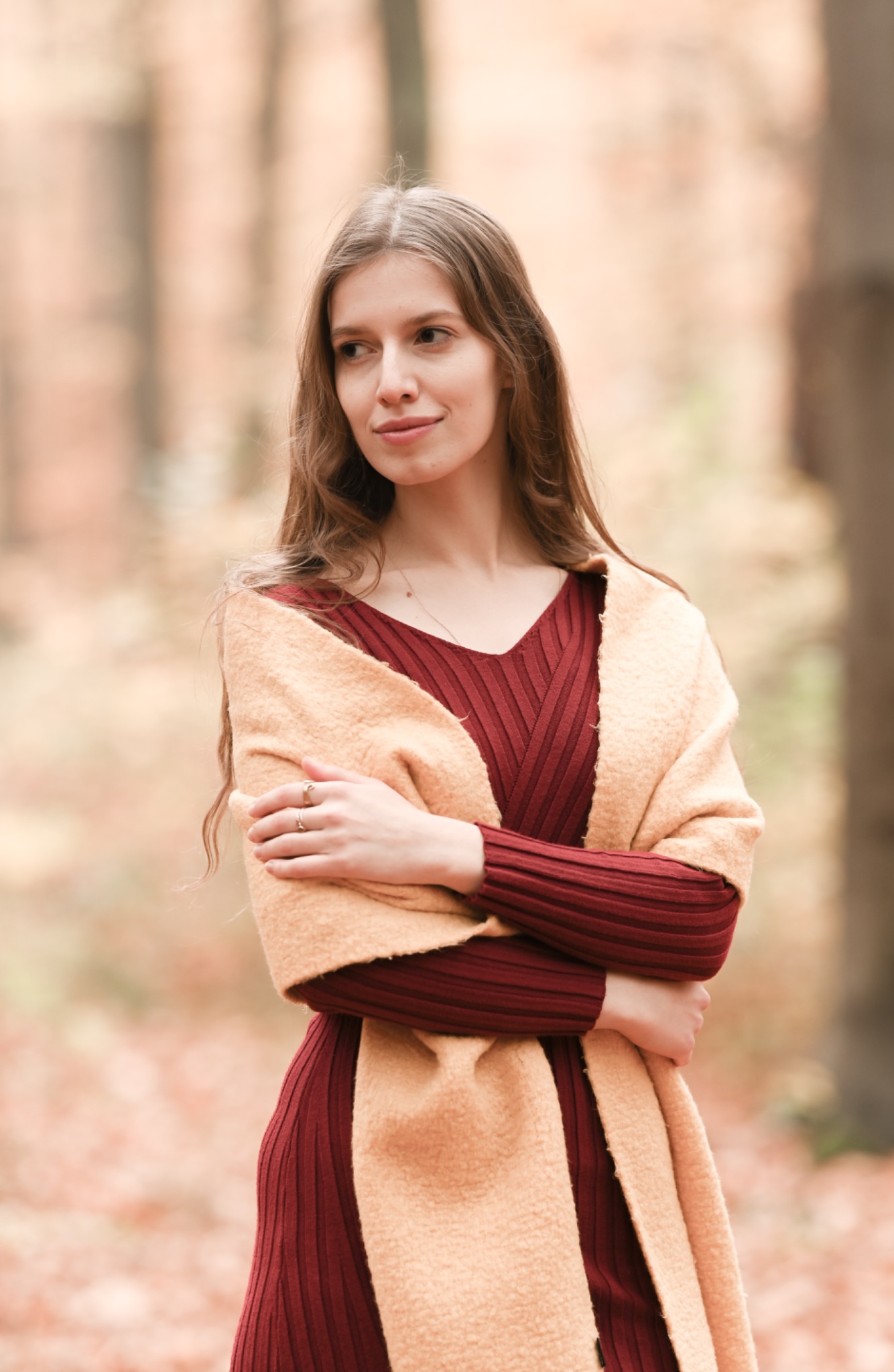 Portret kobiety w bordowej sukience i beżowym szalu na tle jesiennego lasu, z rękami skrzyżowanymi na piersiach i złotymi pierścionkami na palcach.
