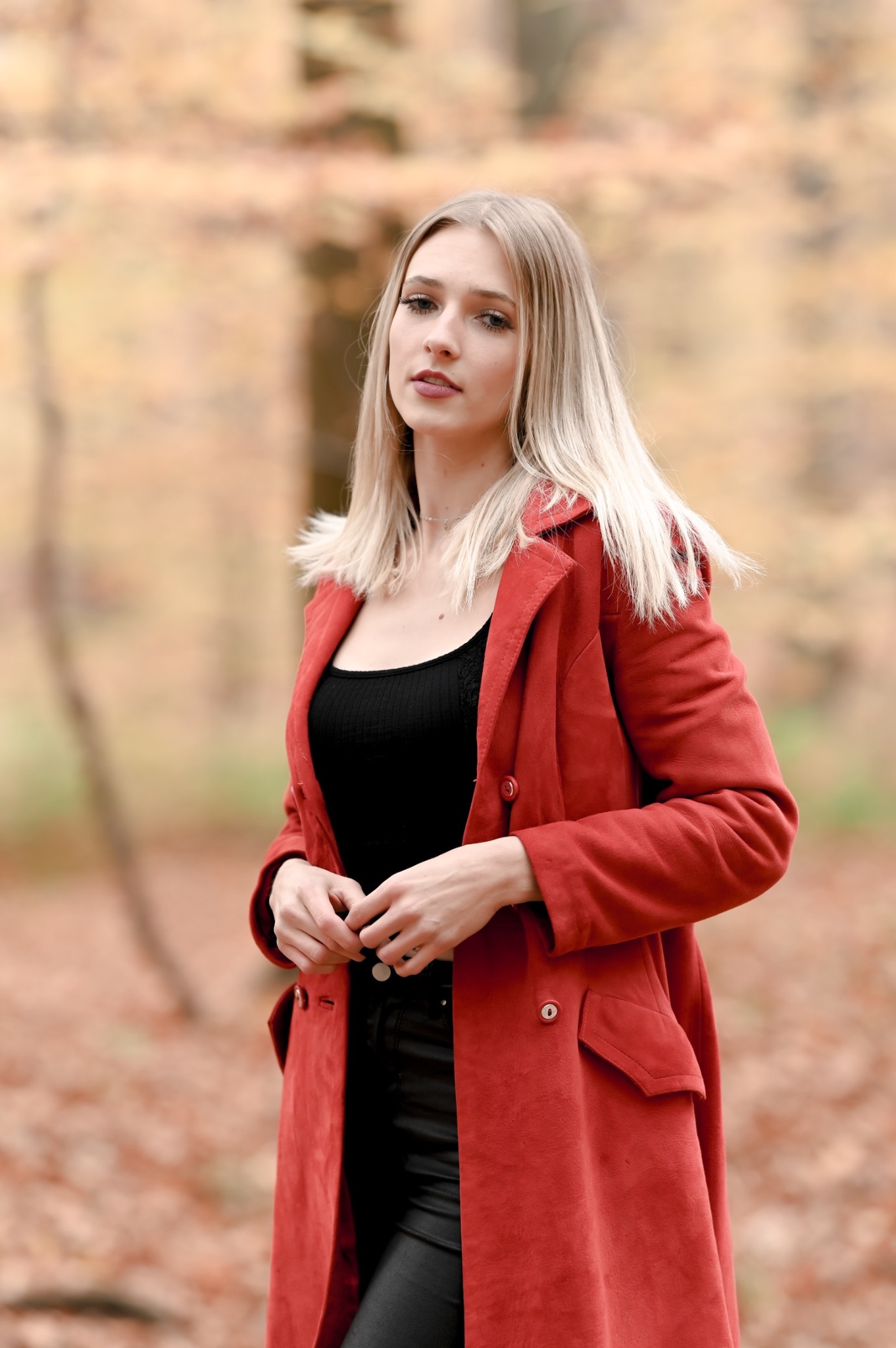 Portret kobiety w czerwonym płaszczu na tle jesiennego lasu, z delikatnym uśmiechem i spojrzeniem w obiektyw.