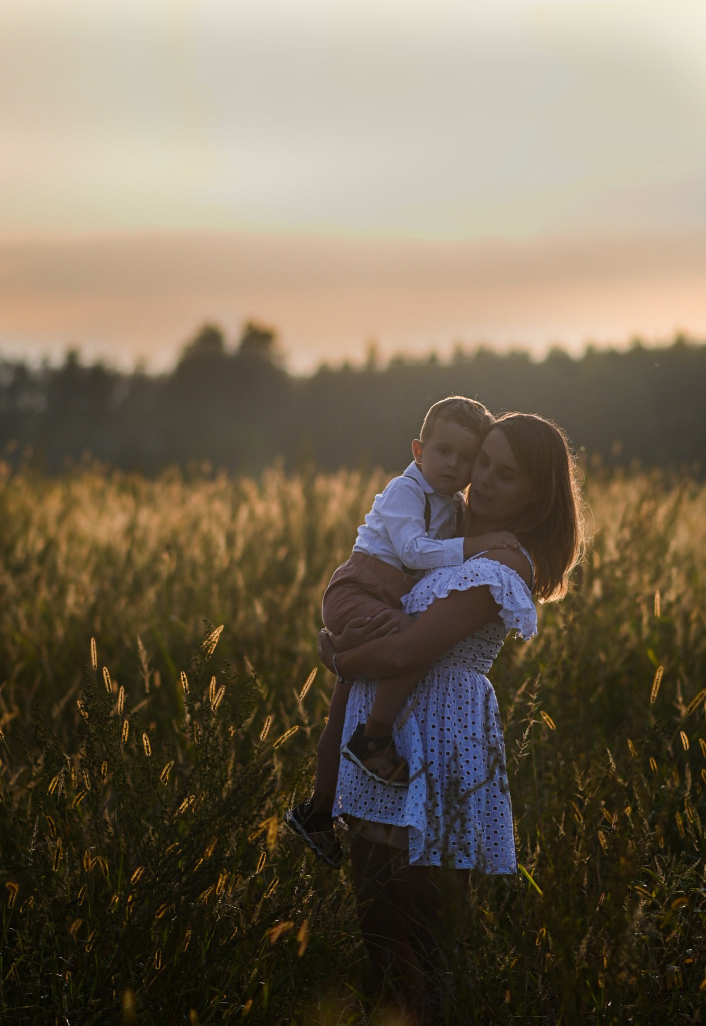 Matka trzymająca syna na tle pola traw w ciepłym świetle zachodzącego słońca, oboje ubrani w jasne ubrania.