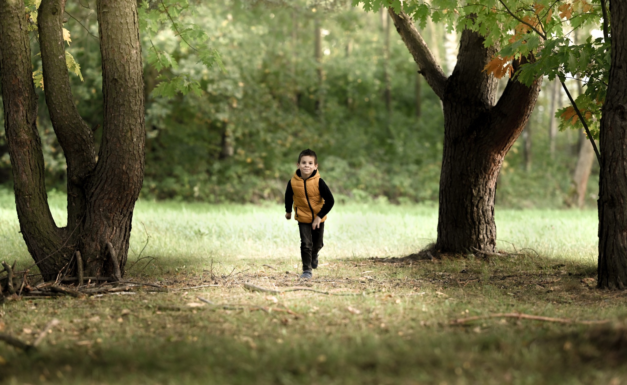 Chłopiec w żółtej kamizelce biegnie przez park, między drzewami, z uśmiechem na twarzy.