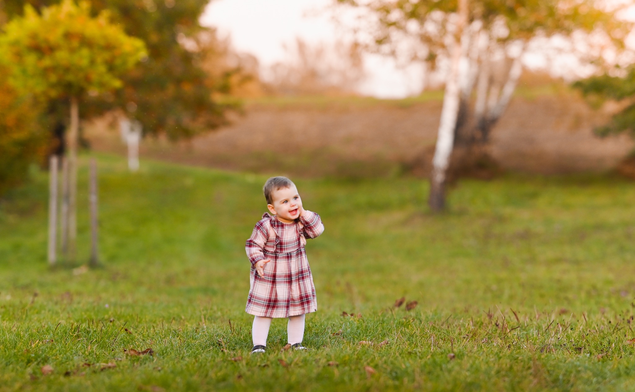 Uśmiechnięta dziewczynka w kraciastej sukience stoi na trawie w parku, z drzewami w tle i delikatnym, rozproszonym światłem.