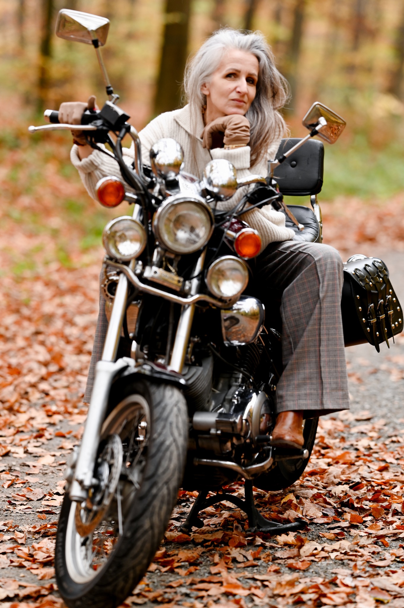 Kobieta w brązowych rękawiczkach i tweedowych spodniach siedzi na czarnym motocyklu na ścieżce pokrytej opadłymi liśćmi, z drzewami w tle.