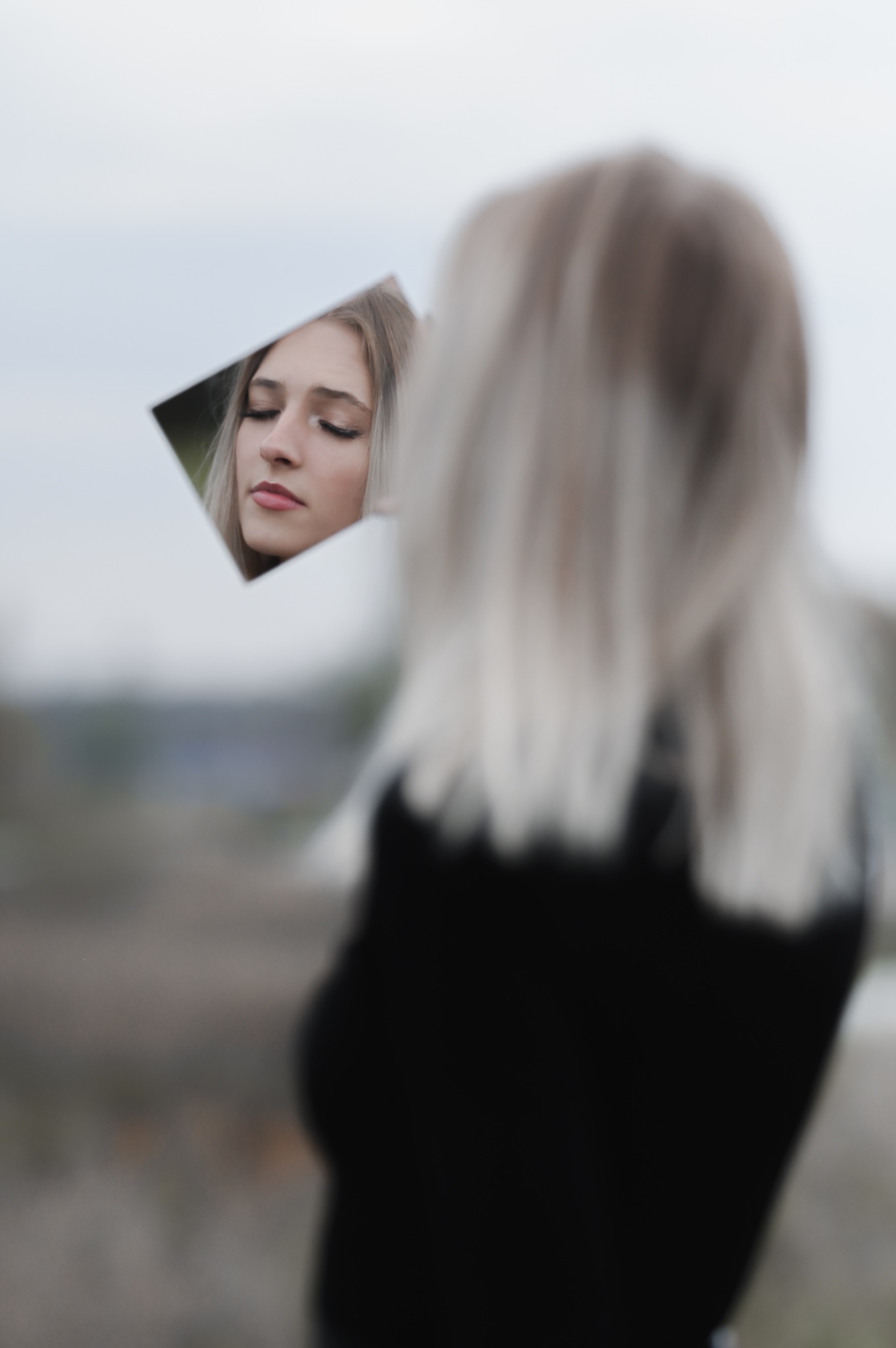Portret kobiety z blond włosami, której twarz odbija się w małym lustrze trzymanym w dłoni, z rozmytym tłem krajobrazu.