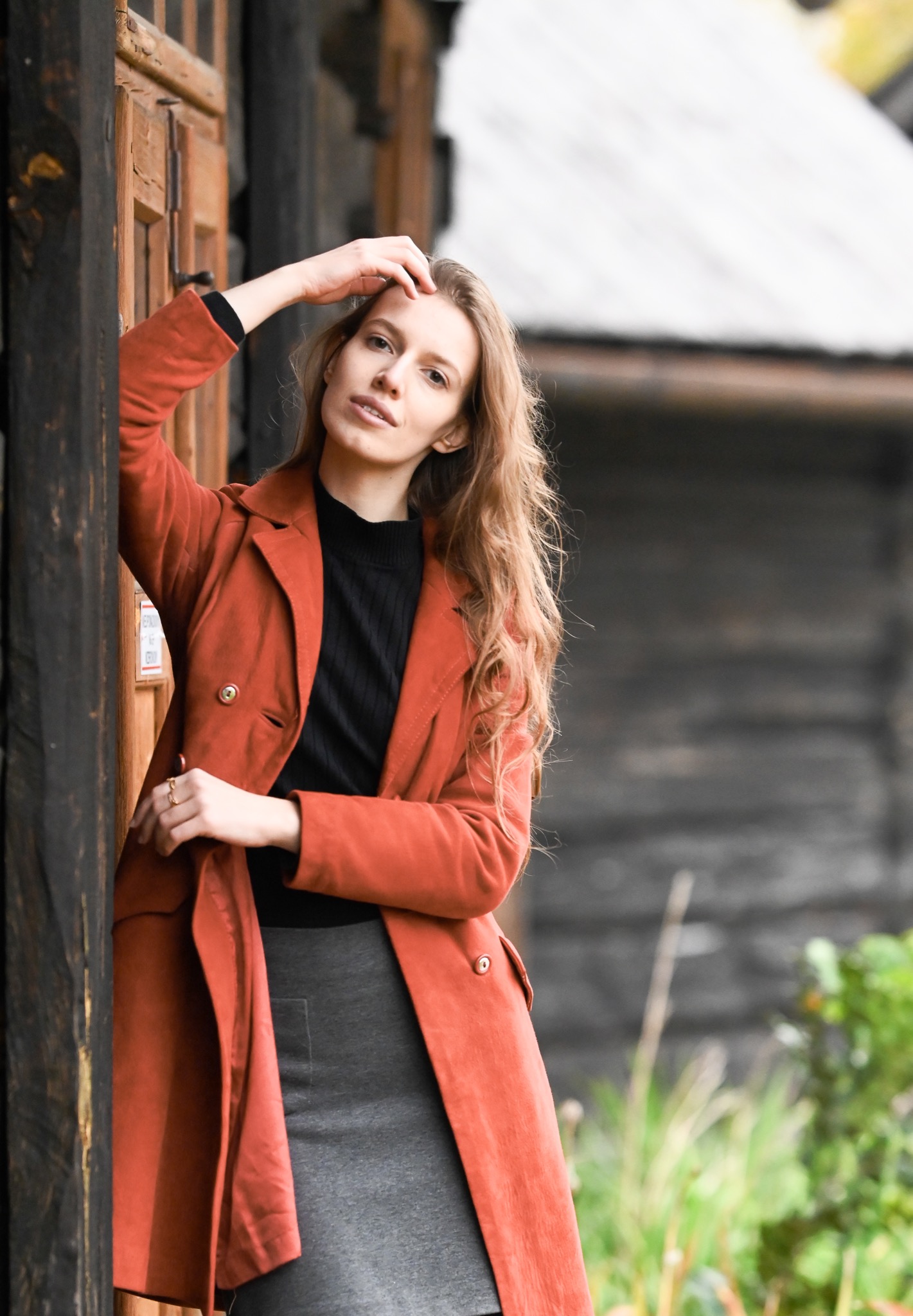 Portret kobiety w pomarańczowym płaszczu, opierającej się o drewnianą ścianę, z rozpuszczonymi włosami i delikatnym uśmiechem.
