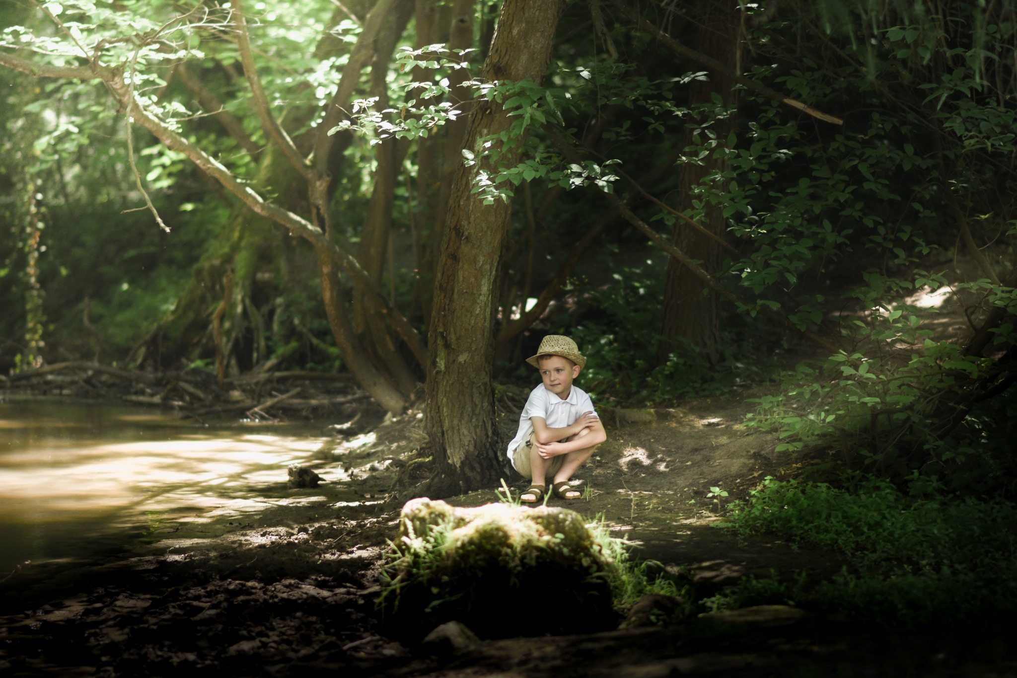 Chłopiec w słomkowym kapeluszu siedzi pod drzewem na skraju lasu, z refleksami światła na wodzie i mchu.