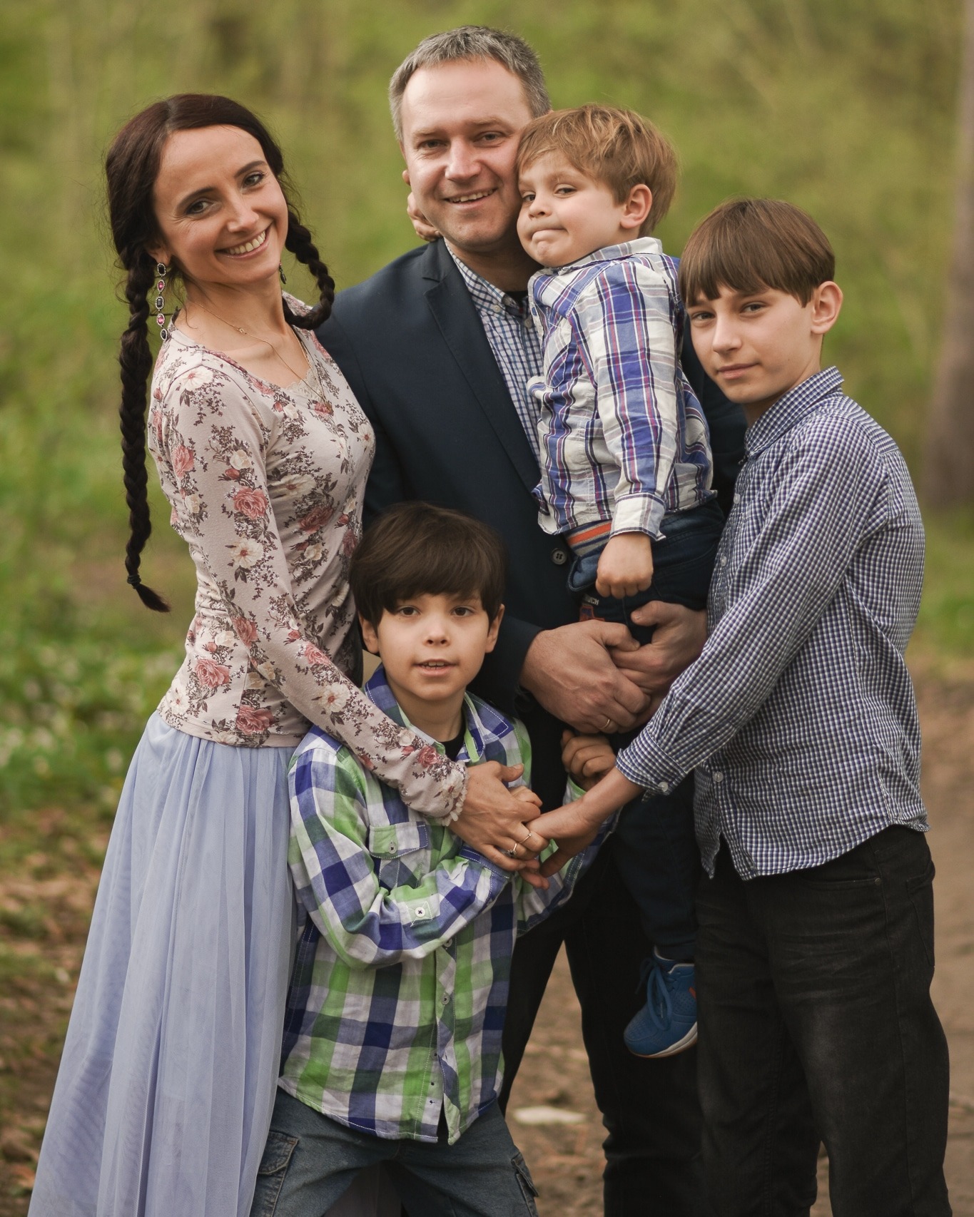 Portret uśmiechniętej rodziny: matka w kwiecistej bluzce i długiej, błękitnej spódnicy, ojciec w marynarce, oraz trzech synów w różnokolorowych koszulach w kratę, na tle zieleni parku.