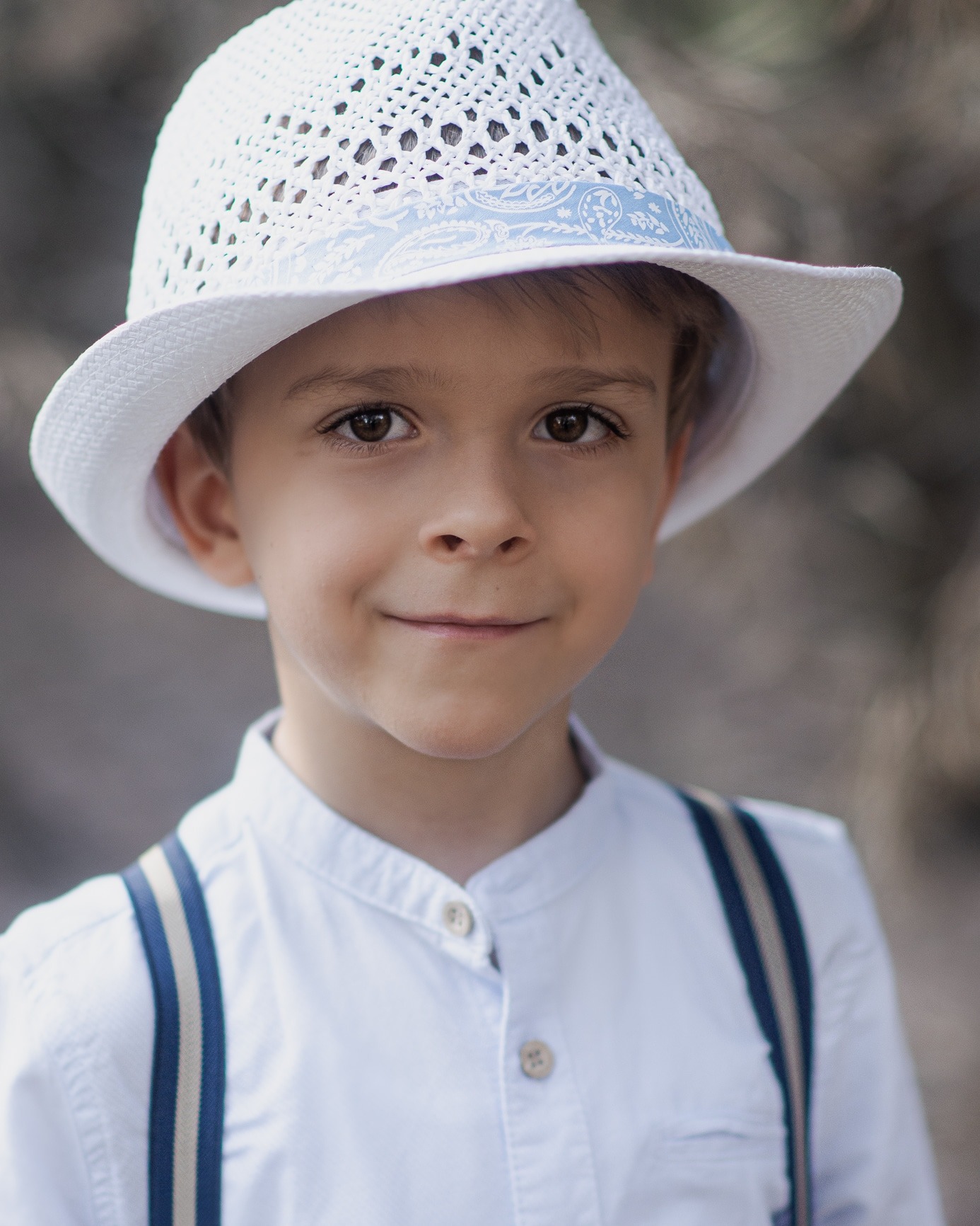 Portret uśmiechniętego chłopca w białym, ażurowym kapeluszu z niebieską wstążką i białej koszuli z szelkami na tle rozmytego otoczenia.