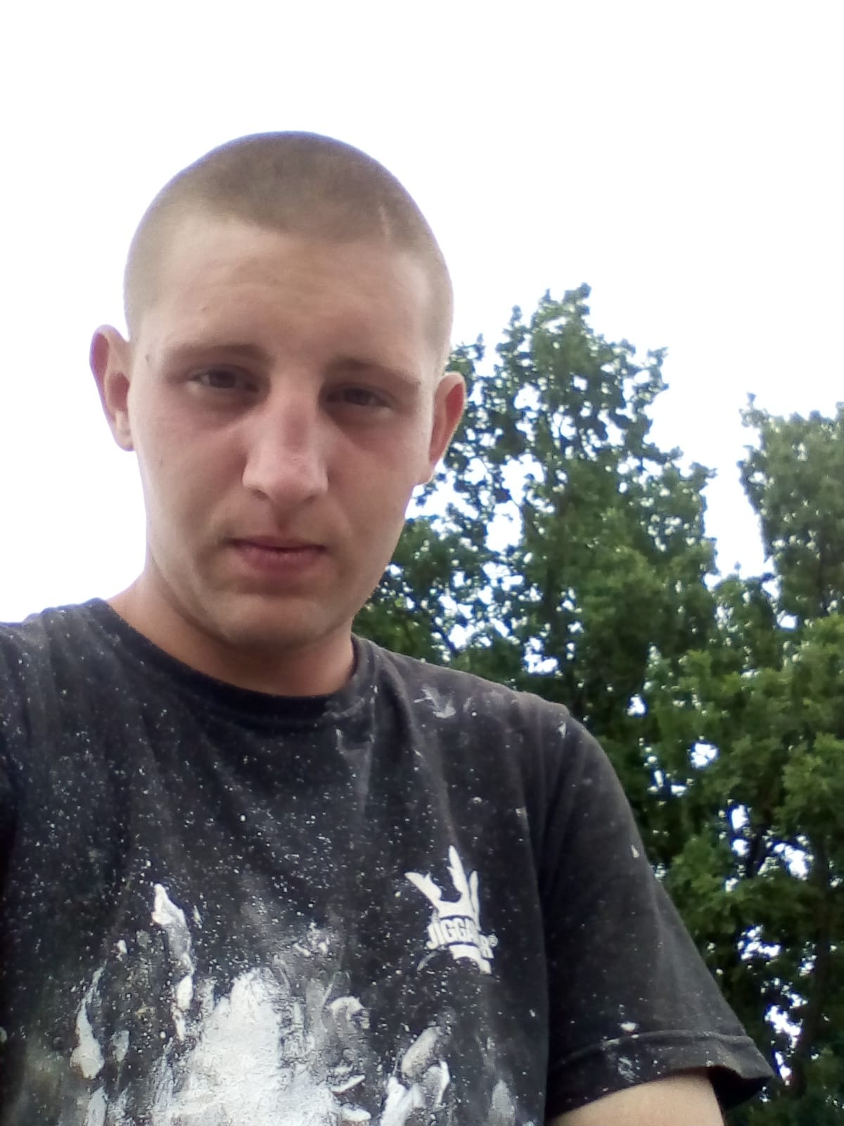 Autoportret mężczyzny z krótkimi włosami, ubranego w czarną koszulkę z białymi plamami farby, z drzewami w tle.