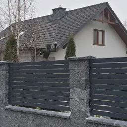 Nowoczesne ogrodzenie z poziomych, grafitowych paneli na niskiej podmurówce z szarej kostki, z filarami z szarego kamienia łupanego, na tle domów jednorodzinnych.