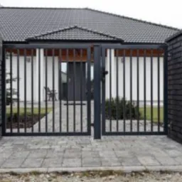 Nowoczesna, metalowa brama wjazdowa z pionowymi szczeblami, osadzona między dwoma murowanymi słupkami z ciemnej cegły klinkierowej, na tle białego domu z szarą dachówką.
