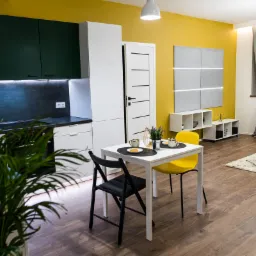 Nowoczesne wnętrze mieszkania z żółtą ścianą, ciemnozielonymi szafkami kuchennymi, białymi meblami i drewnianą podłogą. Stół z czarnym i żółtym krzesłem, nakryty do śniadania.