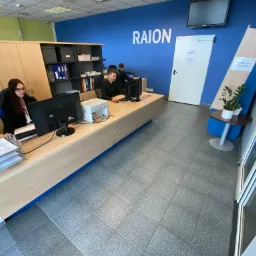 Biuro firmy Raion z pracownikami przy biurkach, komputerami i dokumentami, widoczne logo firmy na ścianie.