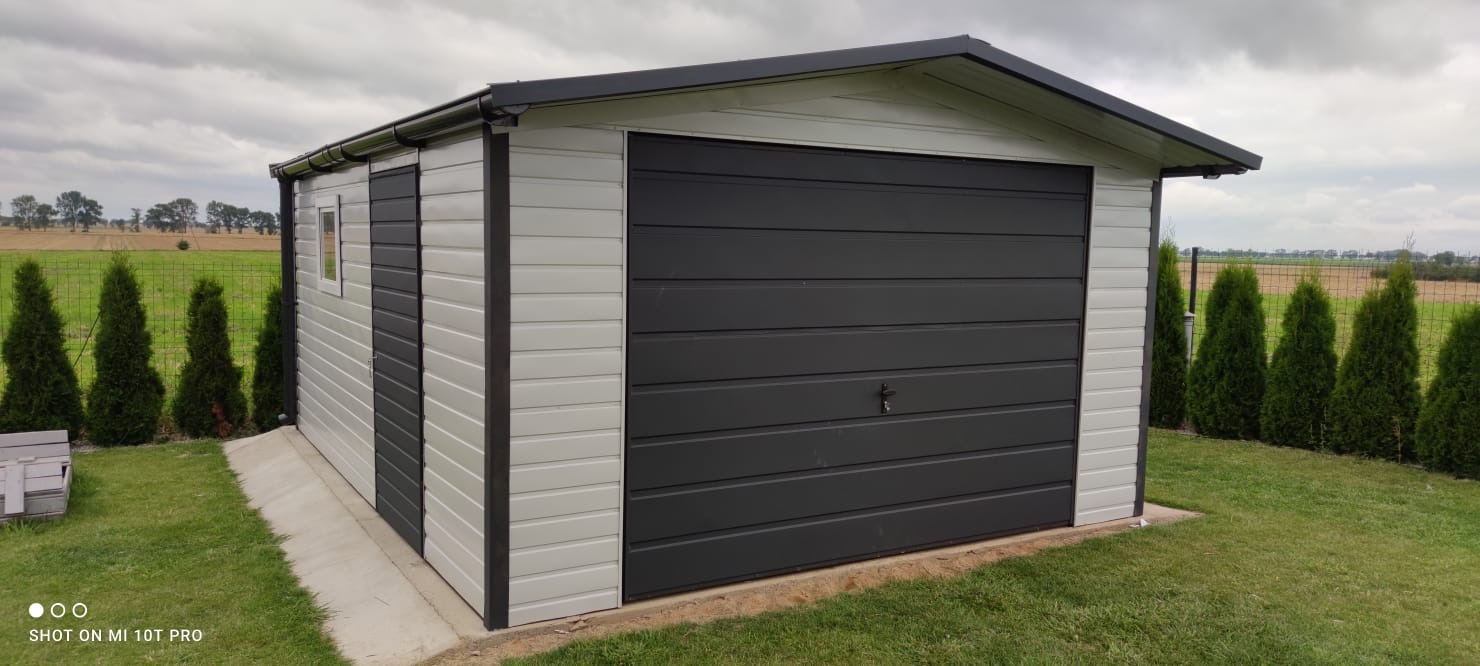 Gotowy, dwukolorowy garaż blaszany z bramą segmentową, otoczony tujami na tle pola uprawnego pod pochmurnym niebem.