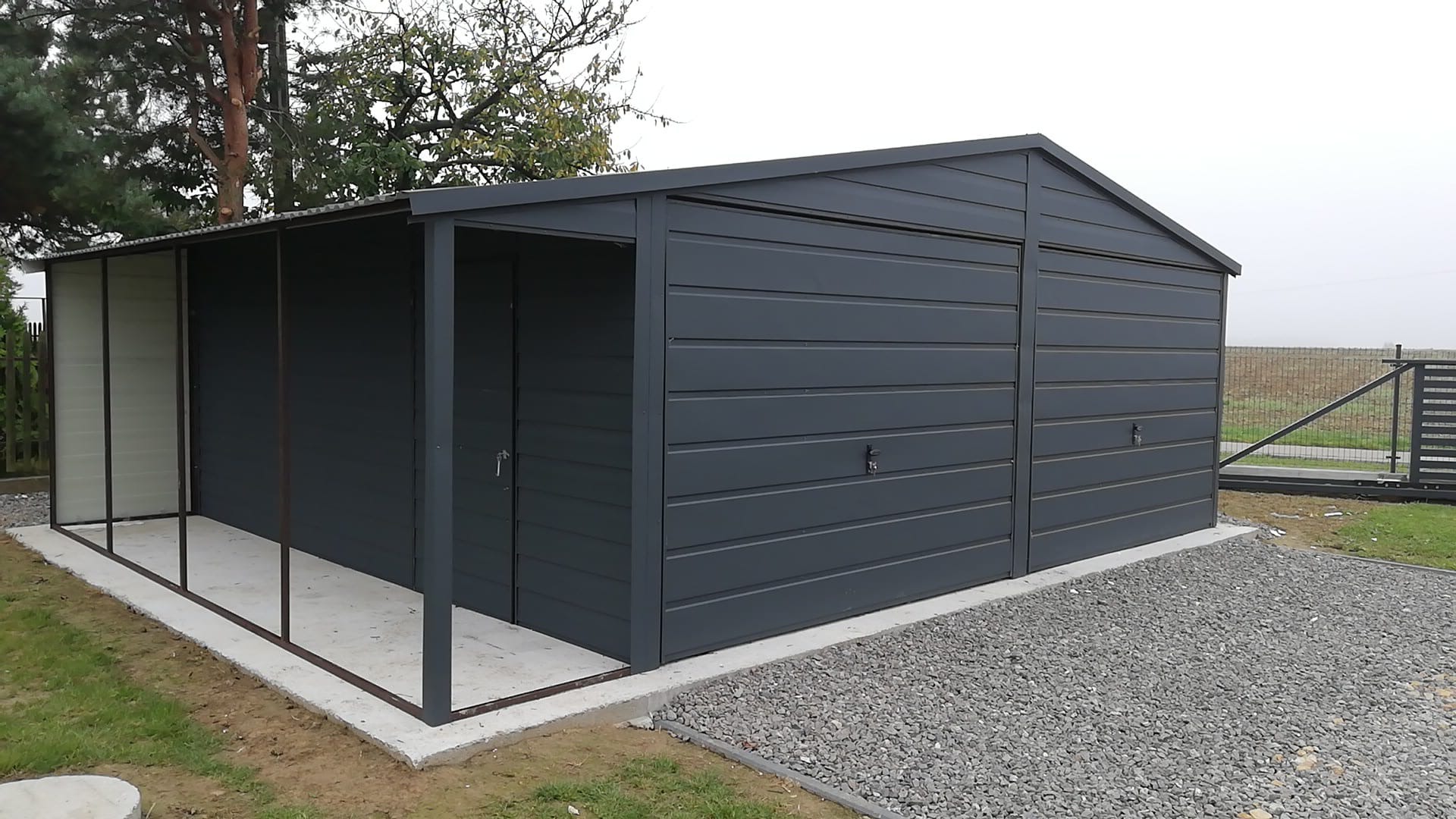 Grafitowy garaż blaszany z dwuspadowym dachem i dobudowaną wiatą, posadowiony na betonowej wylewce z szarym żwirem wokół.
