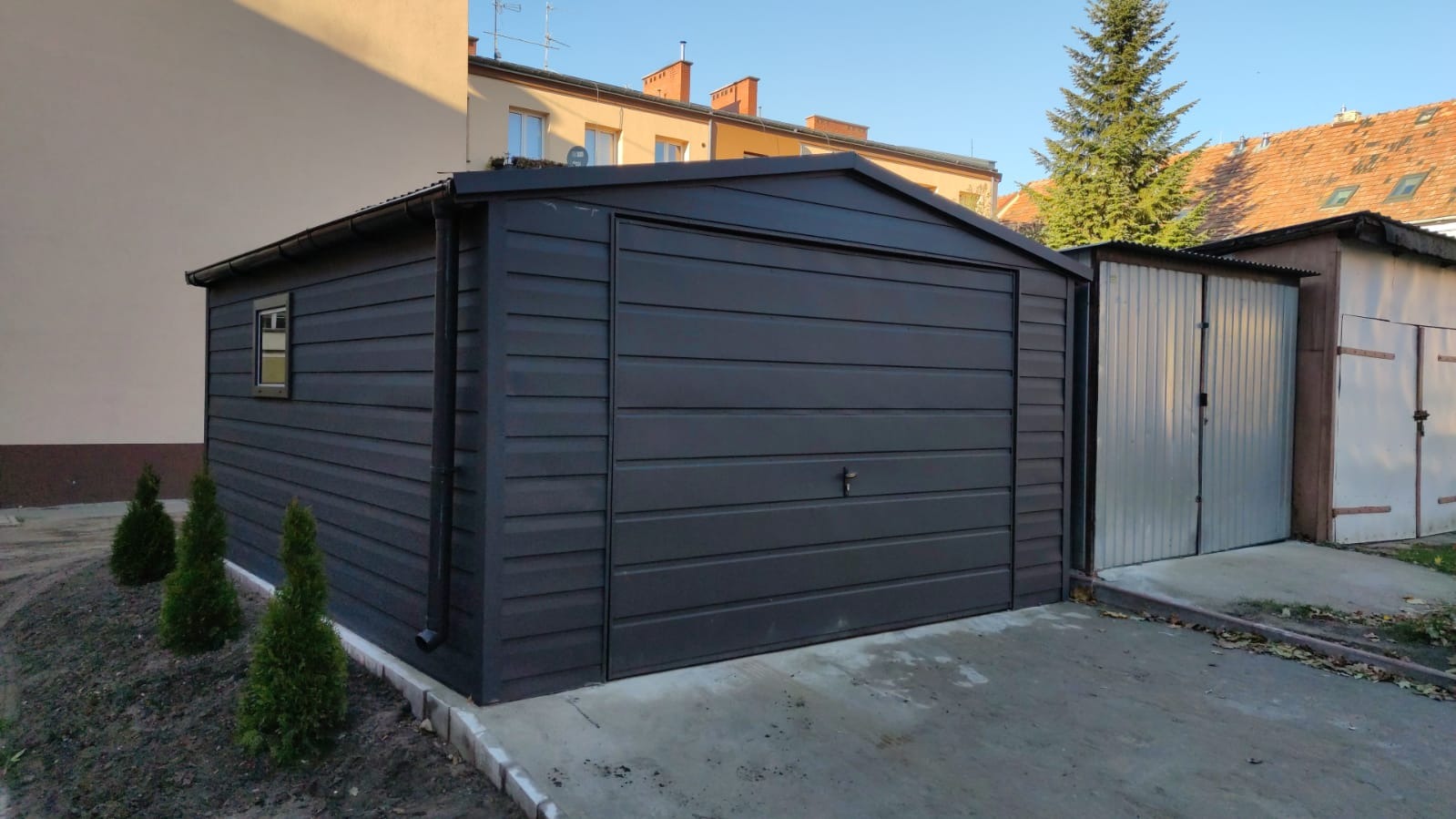 Grafitowy garaż blaszany z oknem, ustawiony na betonowej wylewce, otoczony młodymi tujami i innymi garażami w tle.