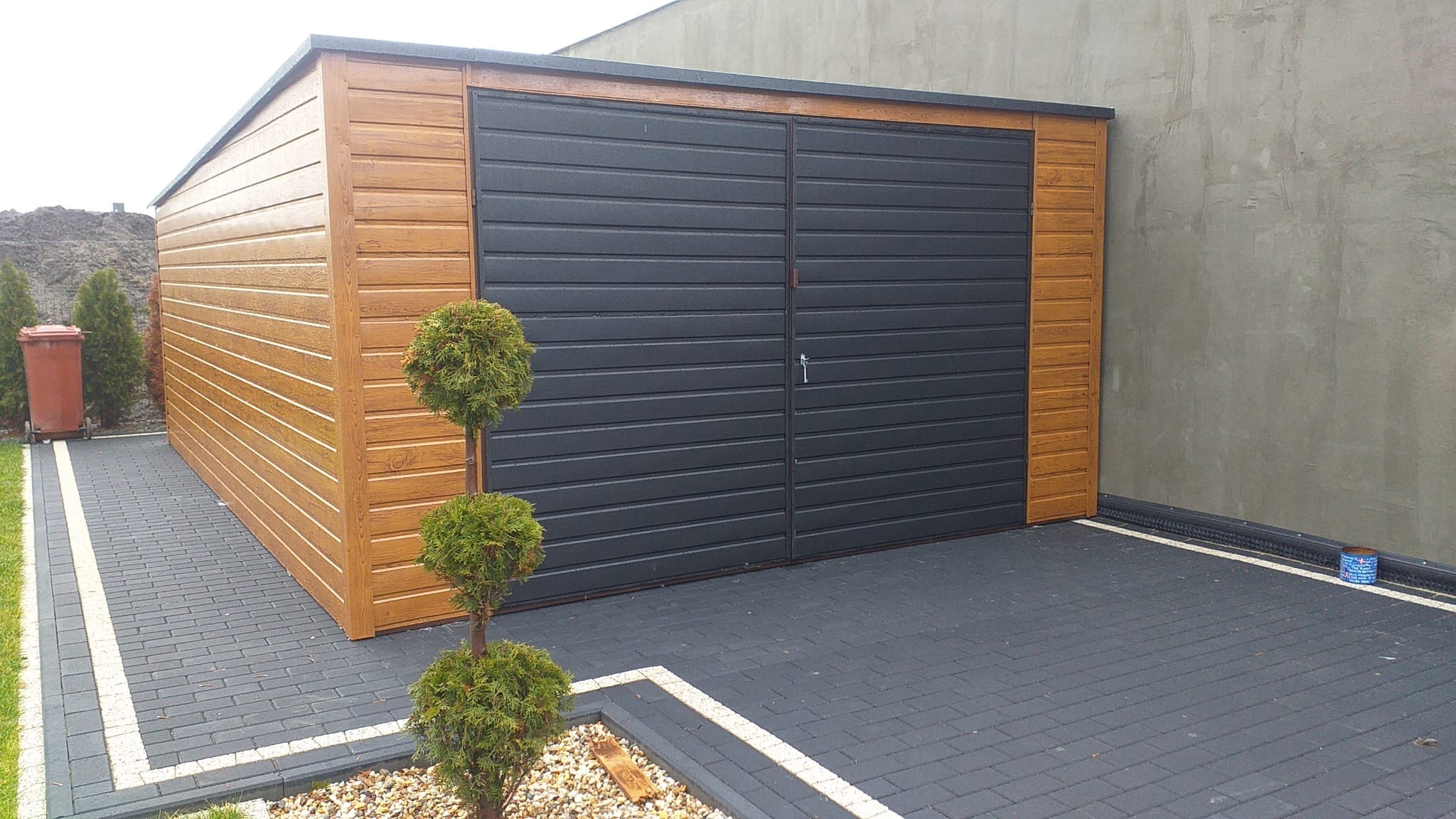 Nowoczesny garaż blaszany z dwuskrzydłowymi, ciemnymi drzwiami, wykończony imitacją drewna, otoczony kostką brukową i ozdobnymi drzewkami.