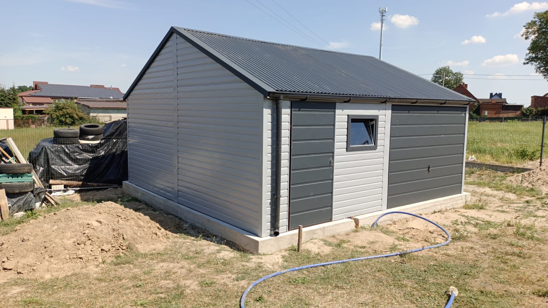 Szary garaż blaszany z dachem dwuspadowym, oknem PCV i bramą uchylną, profil zamknięty poziomy, narożniki i wiatrownice, widoczne opony i narzędzia w tle, teren wokół garażu z ziemią i trawą.