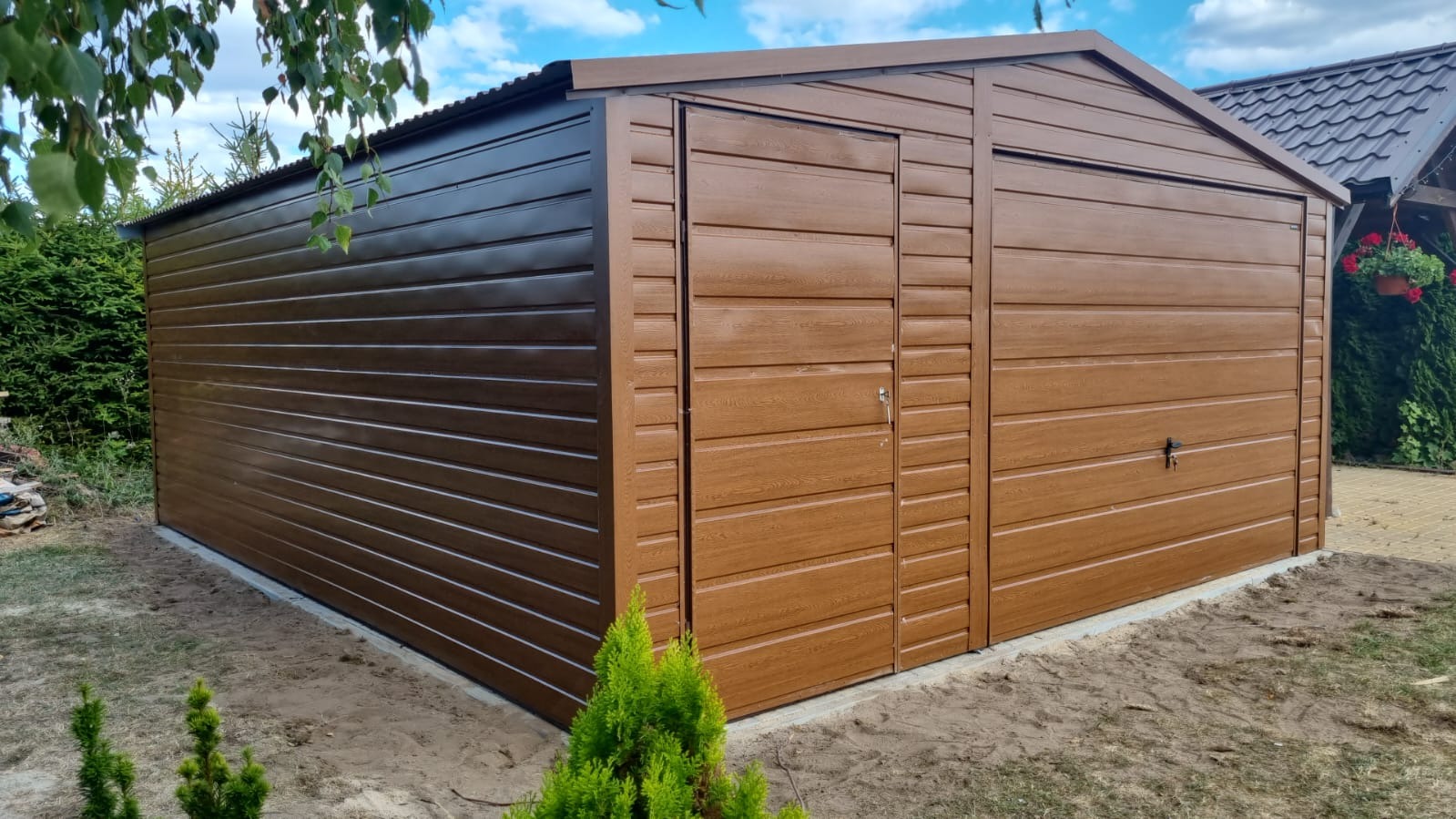 Brązowy garaż blaszany z dachem dwuspadowym, bramą uchylną po prawej i drzwiami wejściowymi po lewej stronie, wykończony w kolorze złotego dębu z poziomym profilem zamkniętym, ustawiony...