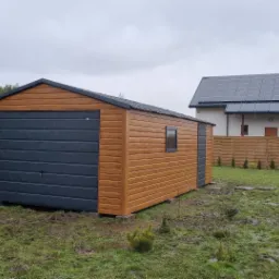 Drewnopodobny garaż z szarą bramą segmentową i małym oknem, ustawiony na betonowych bloczkach na trawniku, w tle dom z panelami słonecznymi i drewniany płot.