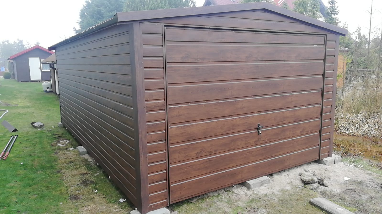 Brązowy, gotowy garaż blaszany z poziomymi panelami, ustawiony na betonowych bloczkach na trawniku, z widocznymi narzędziami obok.