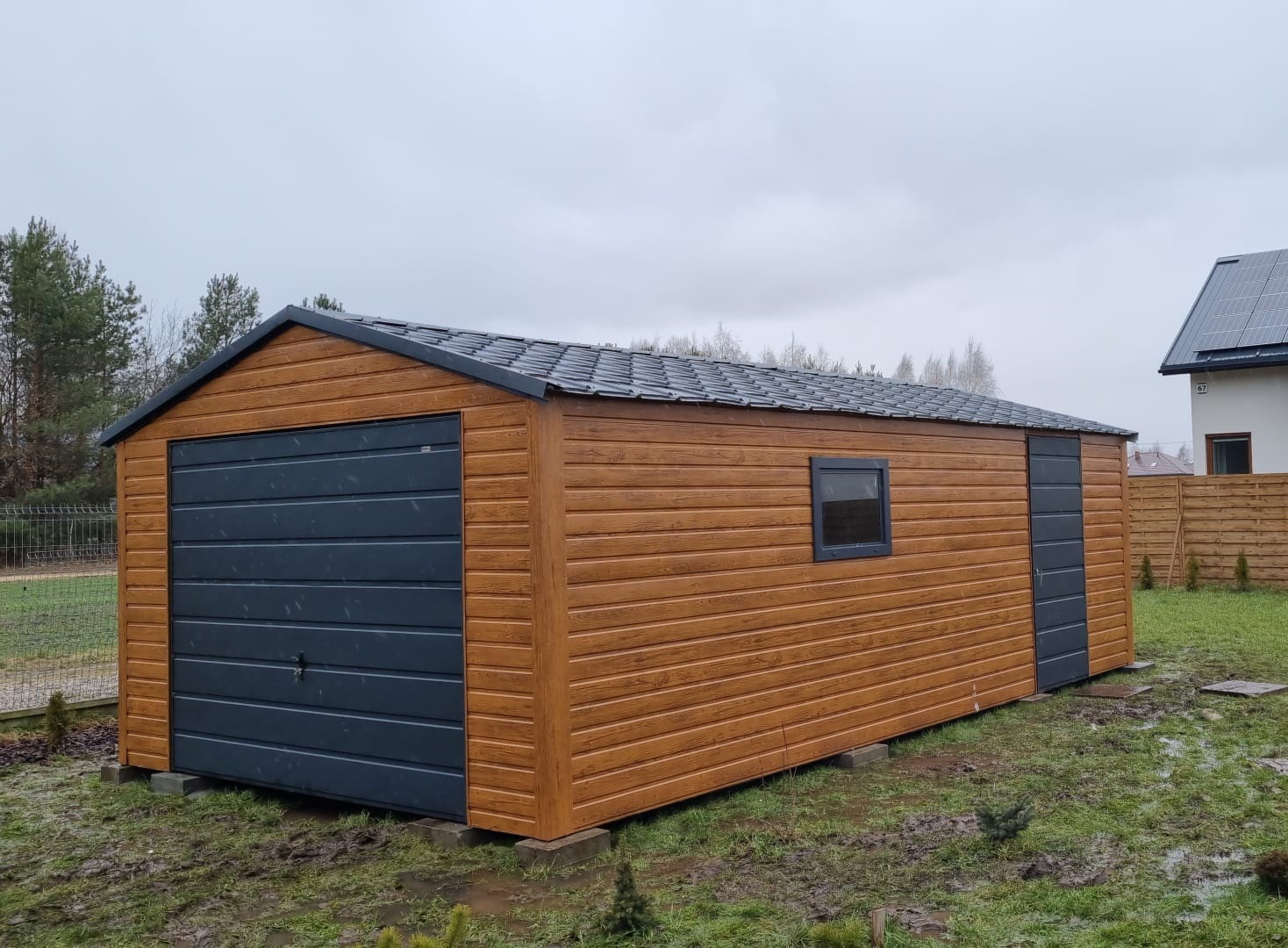 Brązowy garaż blaszany z szarą bramą segmentową i drzwiami bocznymi, widoczny na tle domu z panelami słonecznymi, na trawniku po deszczu.