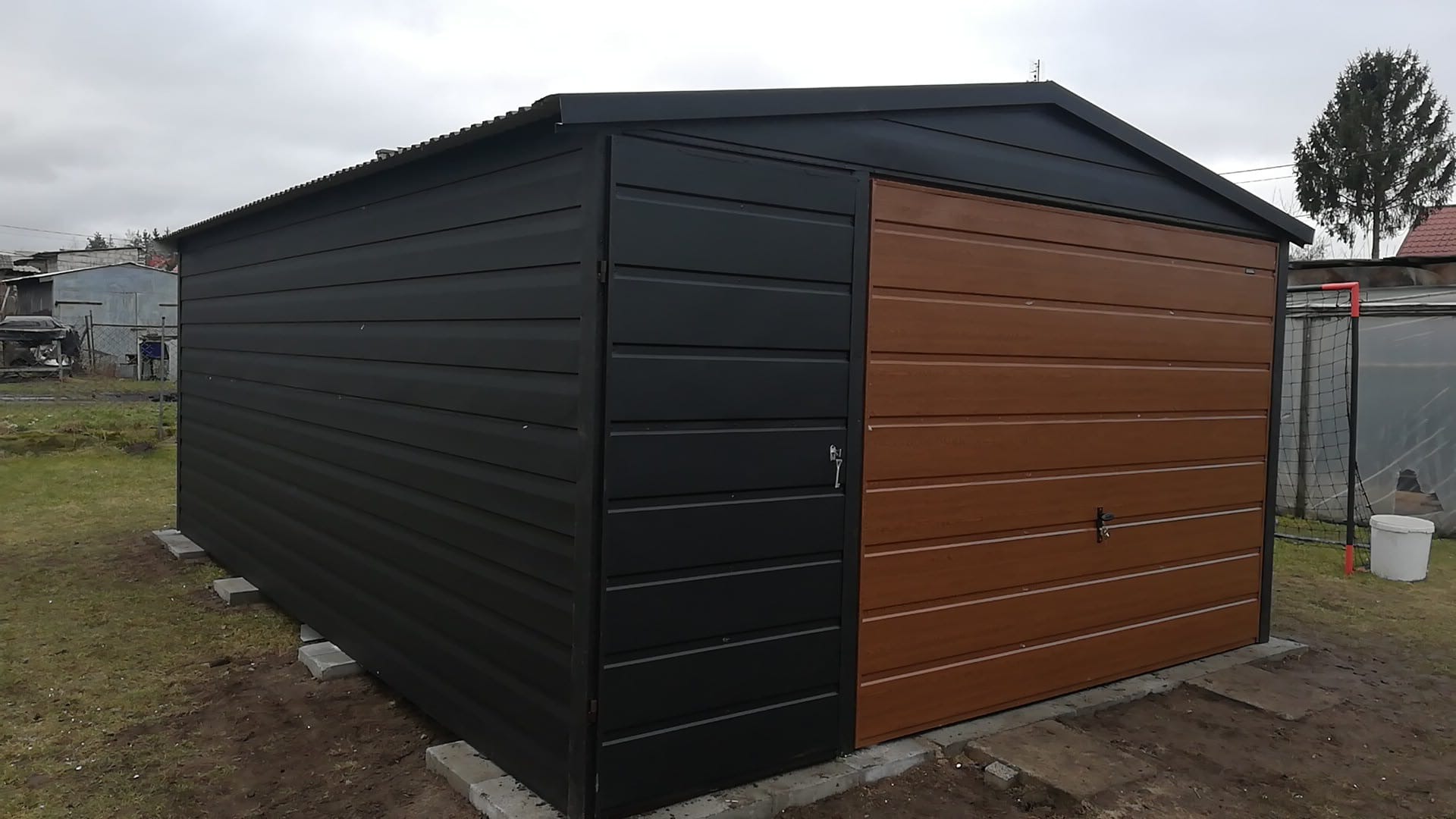 Nowoczesny garaż blaszany z dwukolorową elewacją: czarny bok i brązowe drzwi segmentowe, posadowiony na betonowych bloczkach, z widocznym fragmentem ogrodu w tle.