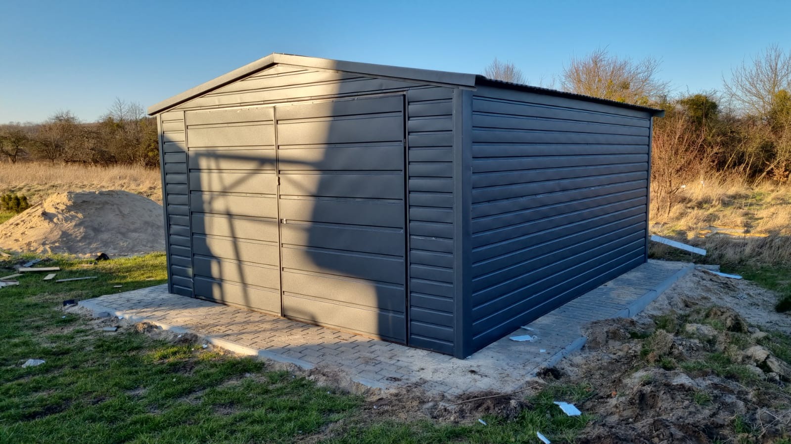 Szary, metalowy budynek gospodarczy z dwuspadowym dachem, posadowiony na fundamencie z kostki brukowej, na tle trawy i drzew.