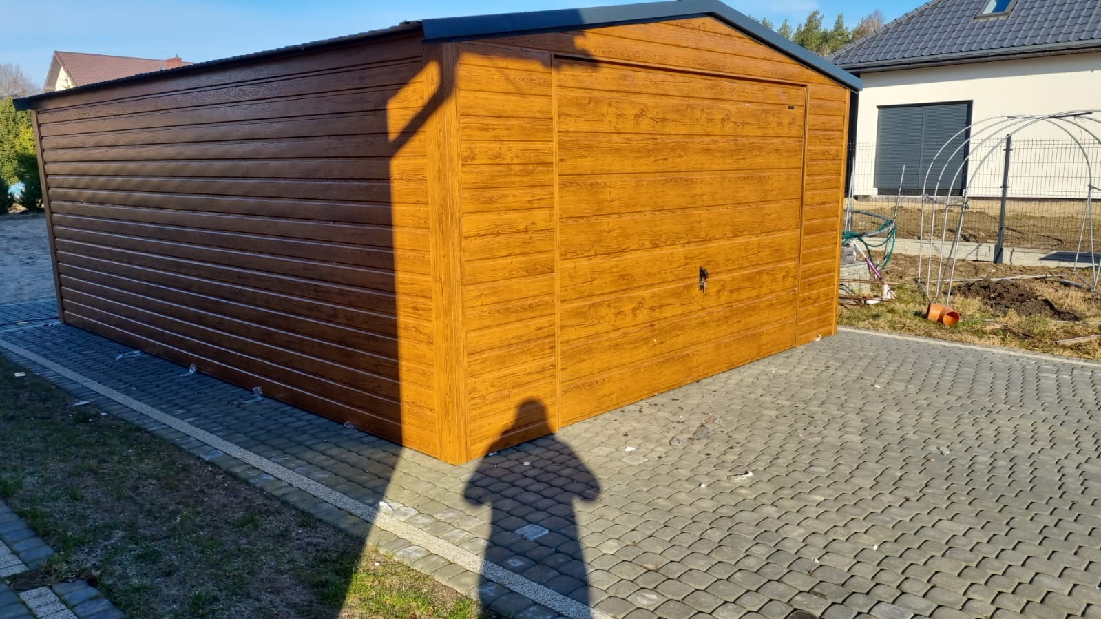 Gotowy, brązowy garaż blaszany z imitacją drewna, stojący na kostce brukowej, z widocznym cieniem osoby robiącej zdjęcie.