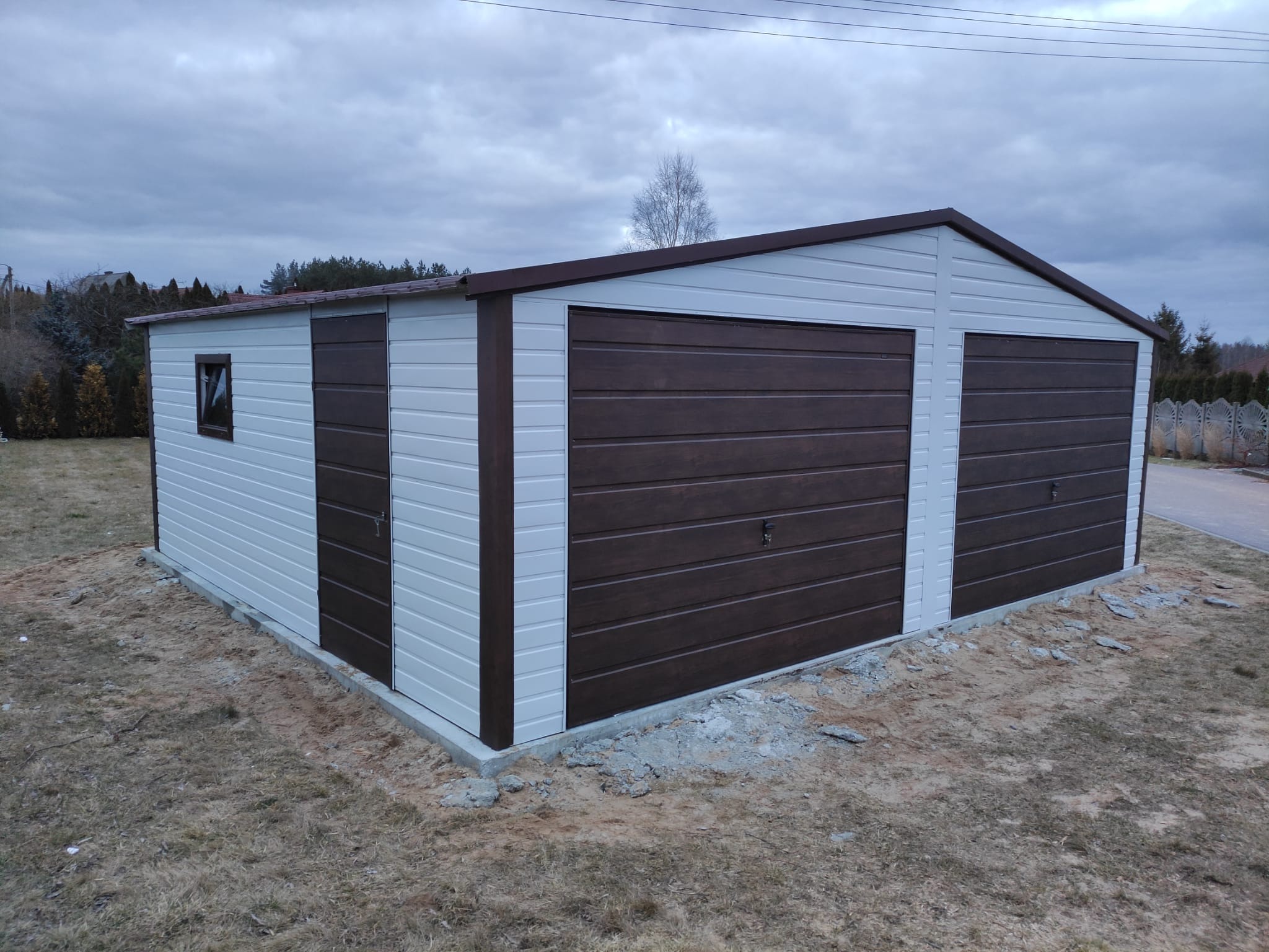 Dwustanowiskowy garaż blaszany z brązowymi bramami segmentowymi i drzwiami bocznymi, widok z zewnątrz na tle pochmurnego nieba.