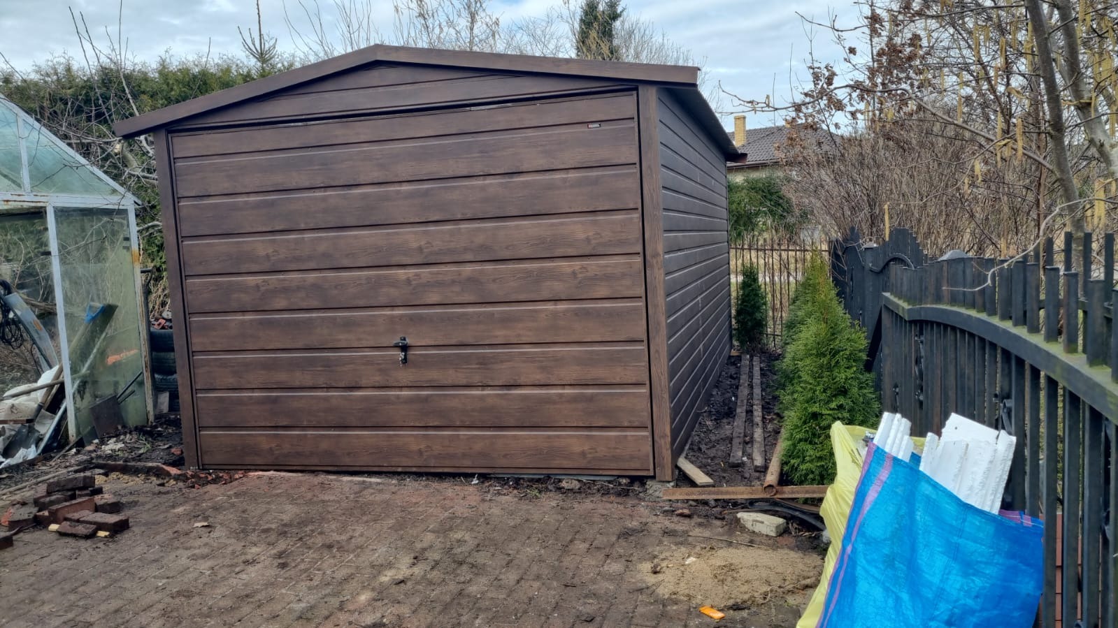 Ciemnobrązowy garaż blaszany z poziomymi panelami, widoczny z zewnątrz na nierównym, ceglanym podłożu, obok starej szklarni i czarnego metalowego ogrodzenia.