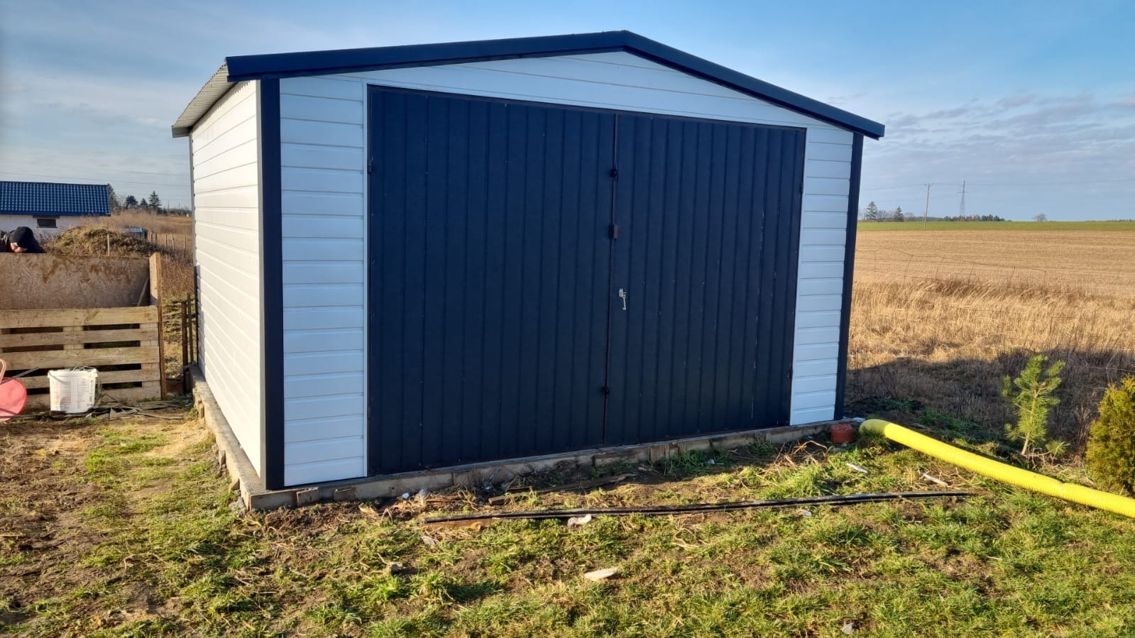 Nowoczesny, biało-grafitowy, mały budynek gospodarczy z dwuskrzydłowymi drzwiami, ustawiony na trawniku z widoczną żółtą rurą odprowadzającą wodę i polami w tle.