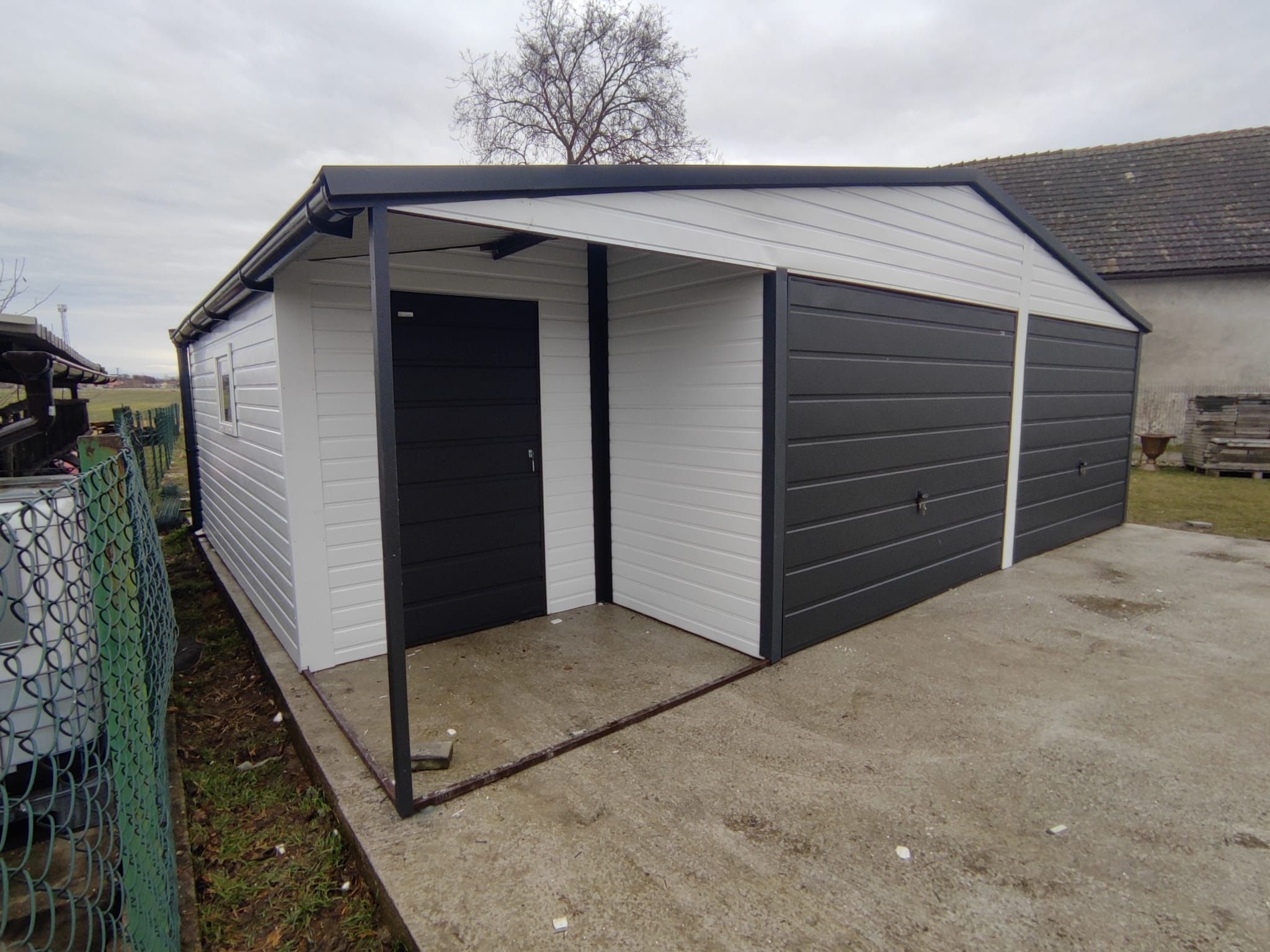 Dwustanowiskowy, blaszany garaż z dodatkowym wejściem, pomalowany na biało-czarno, widoczny na betonowej wylewce w pochmurny dzień.